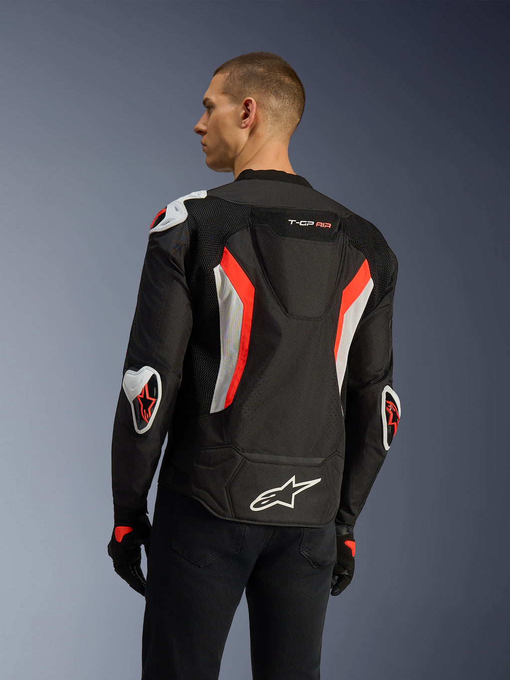Giacca Alpinestars T-GP Air in nero e rosso fluo, vista posteriore a tre quarti su un modello, mette in risalto la gobba aerodinamica, le protezioni esterne Racing GP DFS su spalle e gomiti, i pannelli in mesh traforati al laser e i dettagli bianchi e rosso fluo, giacca da moto in tessuto dal taglio sportivo