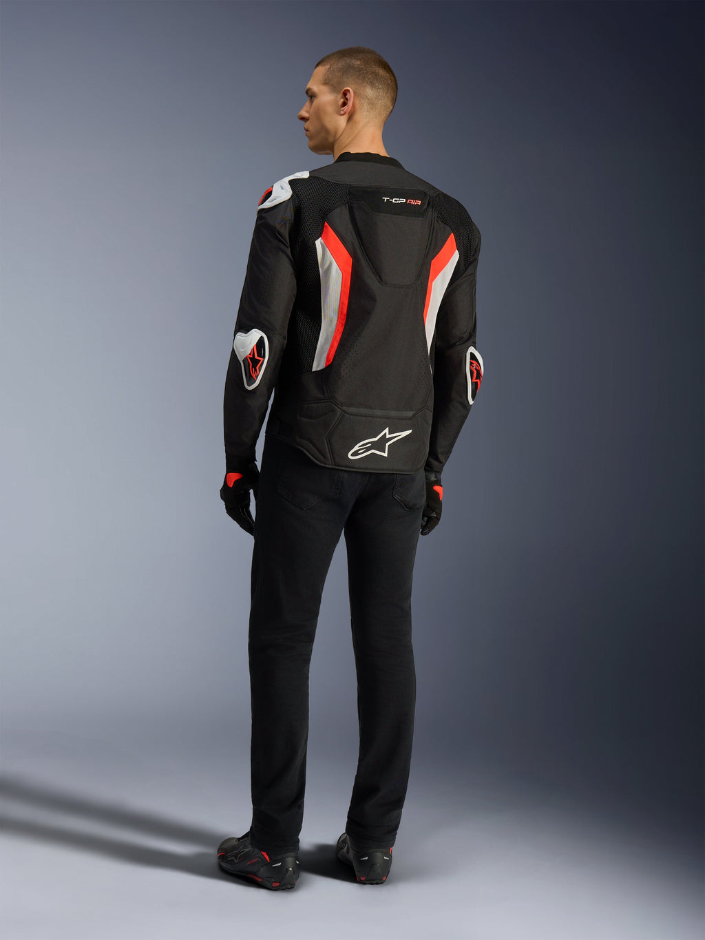 Alpinestars Giacca T-GP Air, giacca da moto Black Red Fluo, vista posteriore su un modello, con pannelli di ventilazione in mesh, gobba aerodinamica e protezioni esterne bianche su gomiti e spalle, indossata con pantaloni neri e scarpe da moto