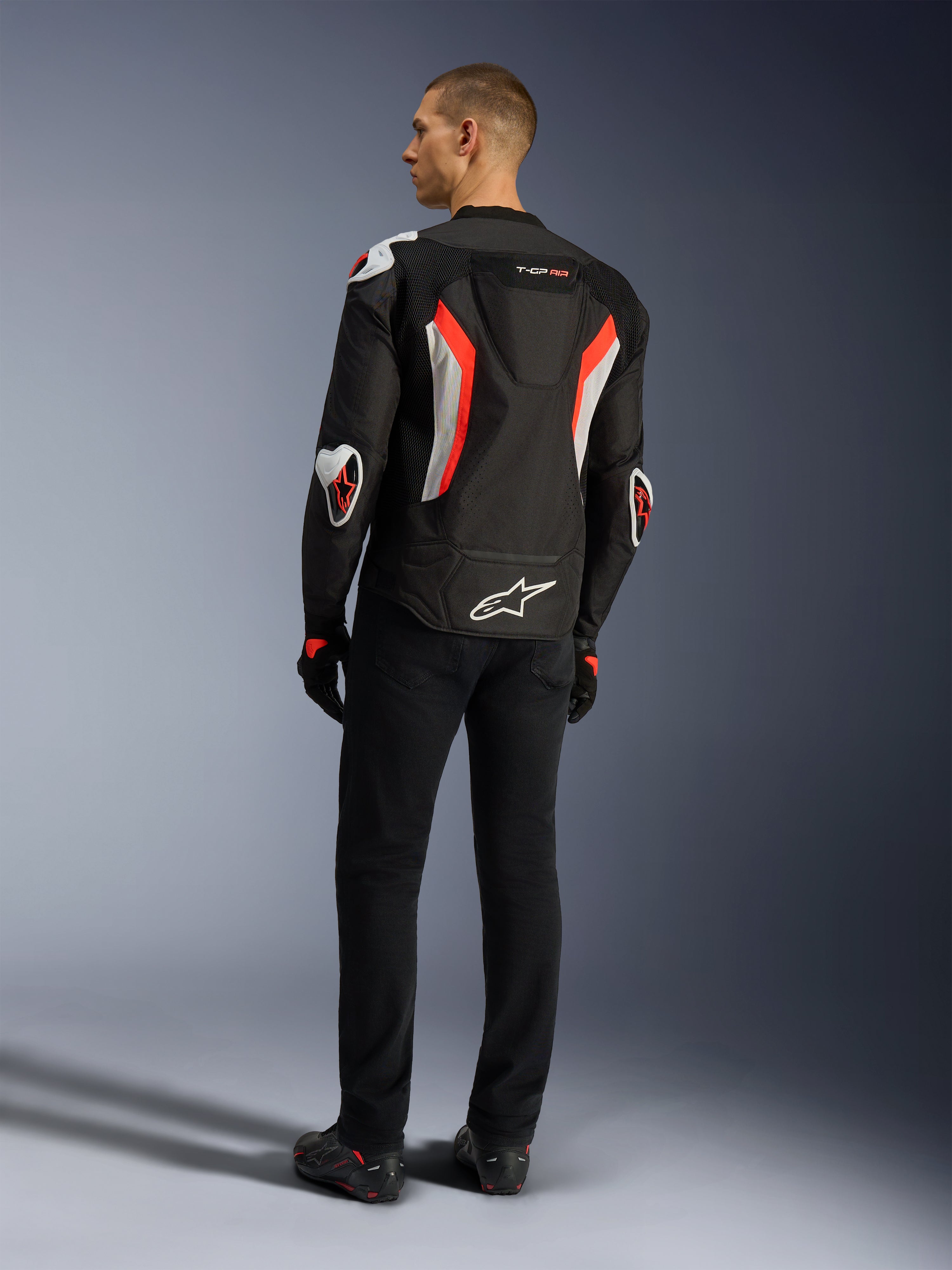 Alpinestars Giacca T-GP Air, giacca da moto Black Red Fluo, vista posteriore su un modello, con pannelli di ventilazione in mesh, gobba aerodinamica e protezioni esterne bianche su gomiti e spalle, indossata con pantaloni neri e scarpe da moto