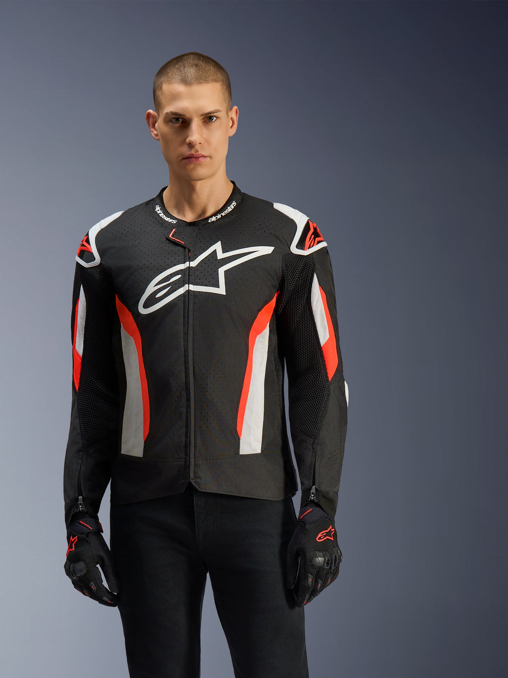 Modello che indossa la giacca Alpinestars T-GP Air, nero con dettagli rosso fluo e bianchi, grande logo Alpinestars sul petto, caratterizzata da pannelli traforati al laser e protezioni per le spalle, indossata con guanti da moto neri, abbigliamento da moto dal taglio sportivo per climi caldi