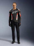 Modello indossa la giacca Alpinestars T-GP Air in Black Red Fluo, giacca da moto in tessuto con accenti bianchi e rosso acceso, grande logo Alpinestars bianco sul petto, con pannelli di ventilazione in mesh, abbinata a pantaloni tecnici neri e guanti da guida, abbigliamento dal taglio sportivo