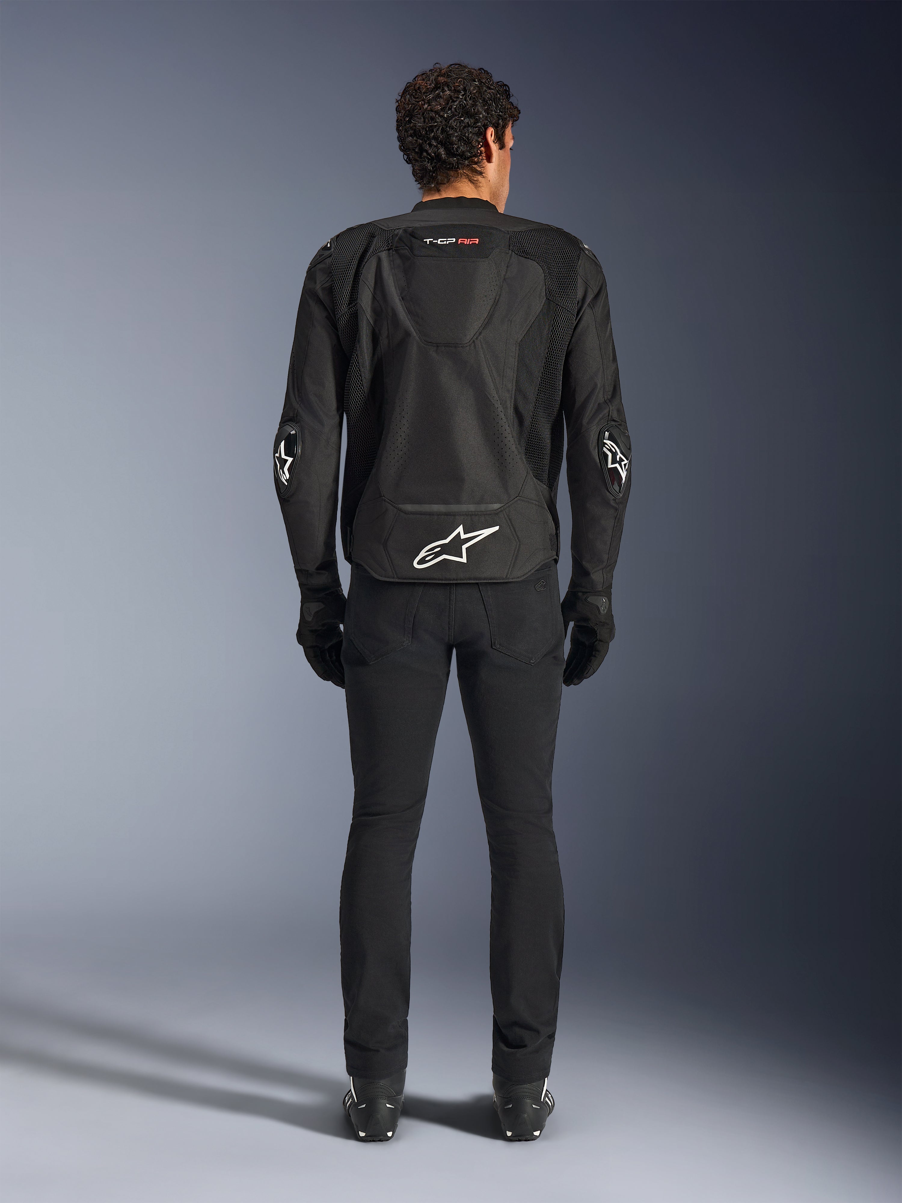 Alpinestars T-GP Air Jacket, giacca da moto in tessuto, nera, vista posteriore di un uomo con pannelli di ventilazione in rete, una gobba aerodinamica e loghi Alpinestars bianchi sulla parte inferiore della schiena e sui gomiti, indossata con pantaloni da guida e guanti neri