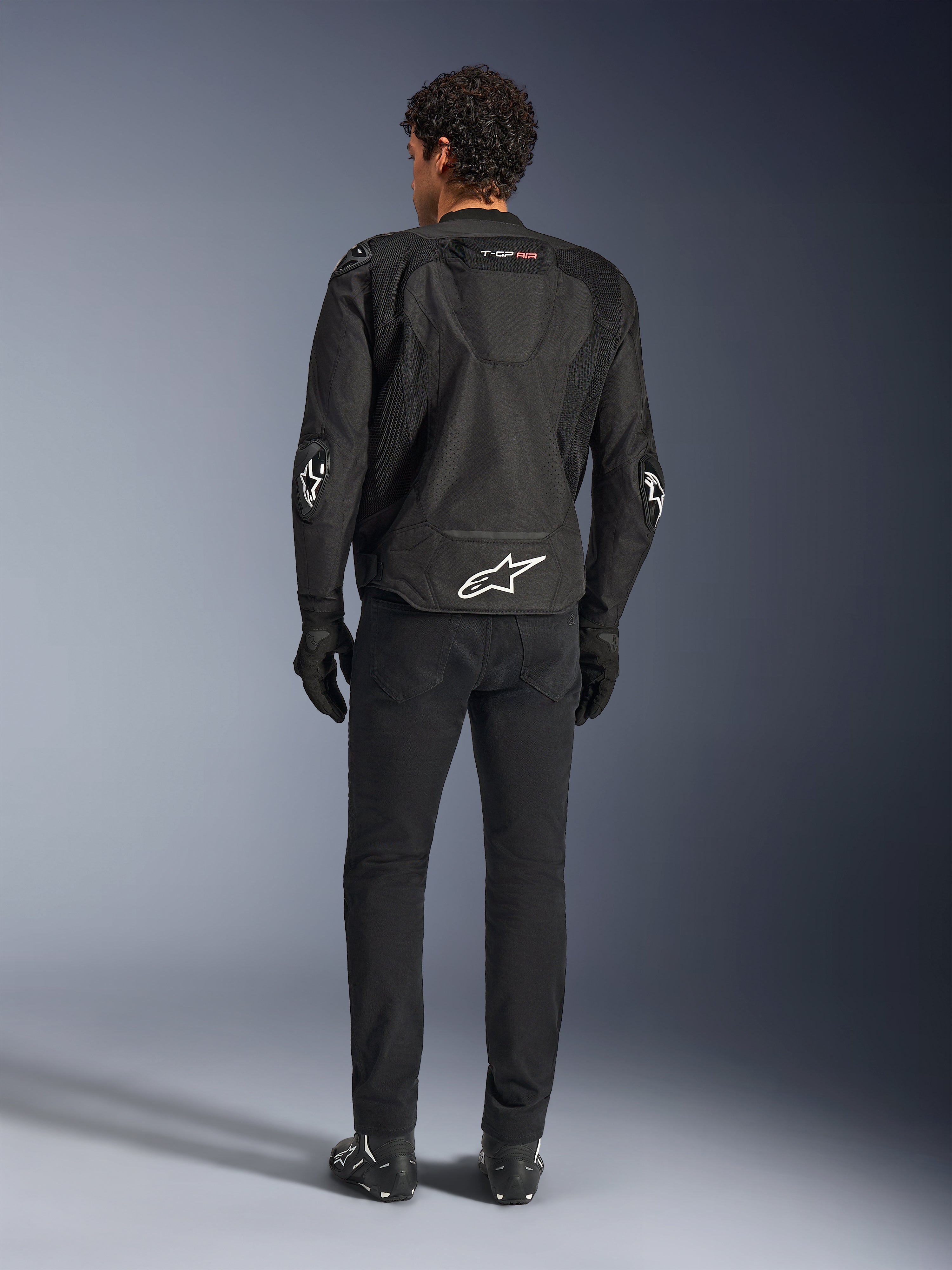 Giacca Alpinestars T-GP Air, nero, vista posteriore su modello, che mostra i pannelli di ventilazione in rete 3D e la gobba aerodinamica, con logo a stella bianco sulla parte bassa della schiena e sui gomiti, indossata con pantaloni e guanti da moto neri, abbigliamento tecnico sportivo.