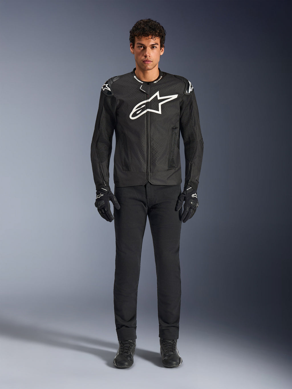 Alpinestars T-GP Air Jacket, giacca da moto in tessuto nero con un grande logo Astars bianco sul petto, indossata da un modello con pantaloni da guida e guanti neri, con pannelli traforati e ventilazione in rete per la guida sportiva