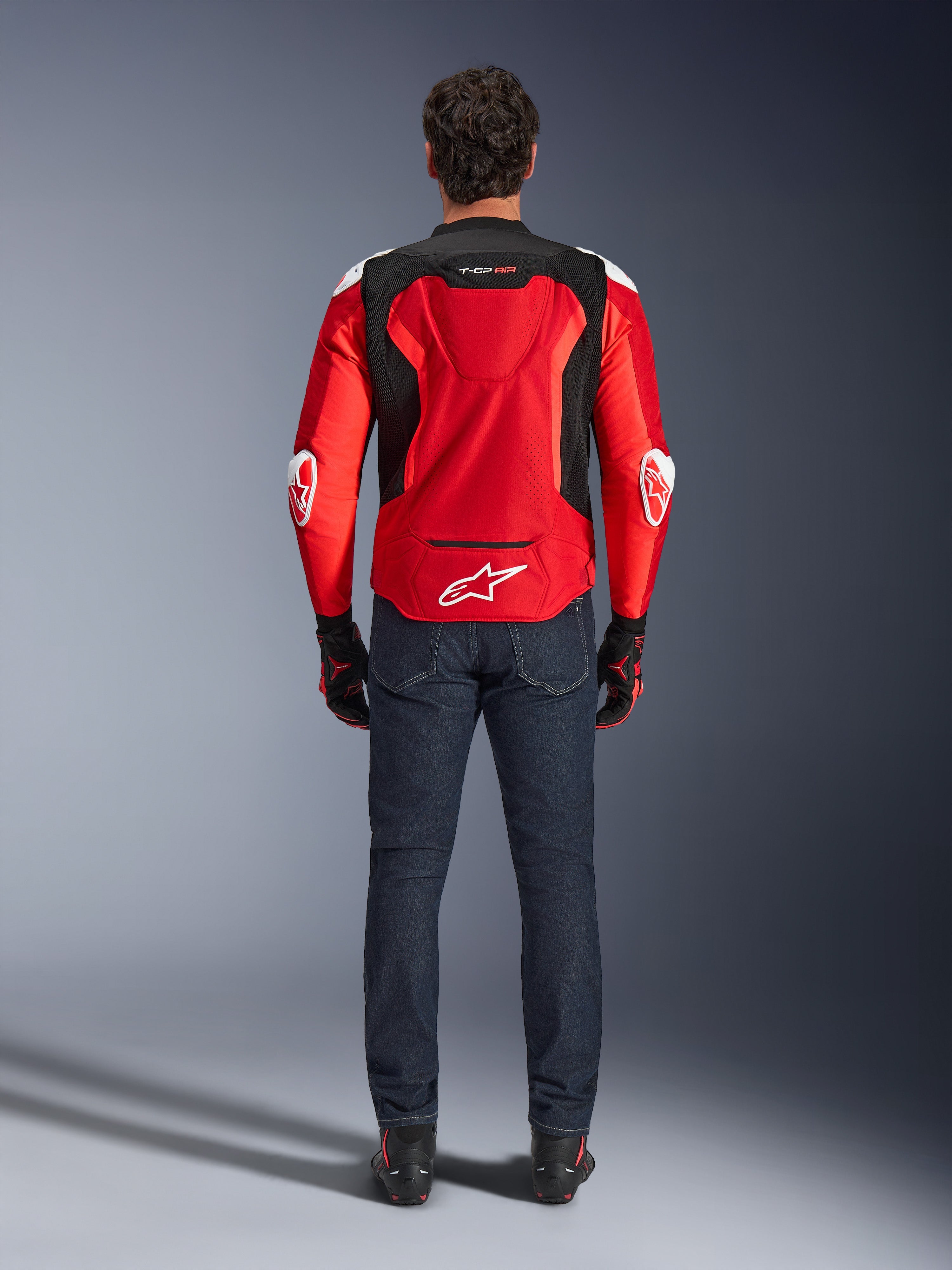 Giacca Alpinestars T-GP Air, vista posteriore, rosso brillante e nero, logo bianco sulla parte inferiore della schiena e marchio 'T-GP AIR', indossata con jeans denim blu scuro e guanti da moto neri e rossi, abbigliamento da moto sportivo