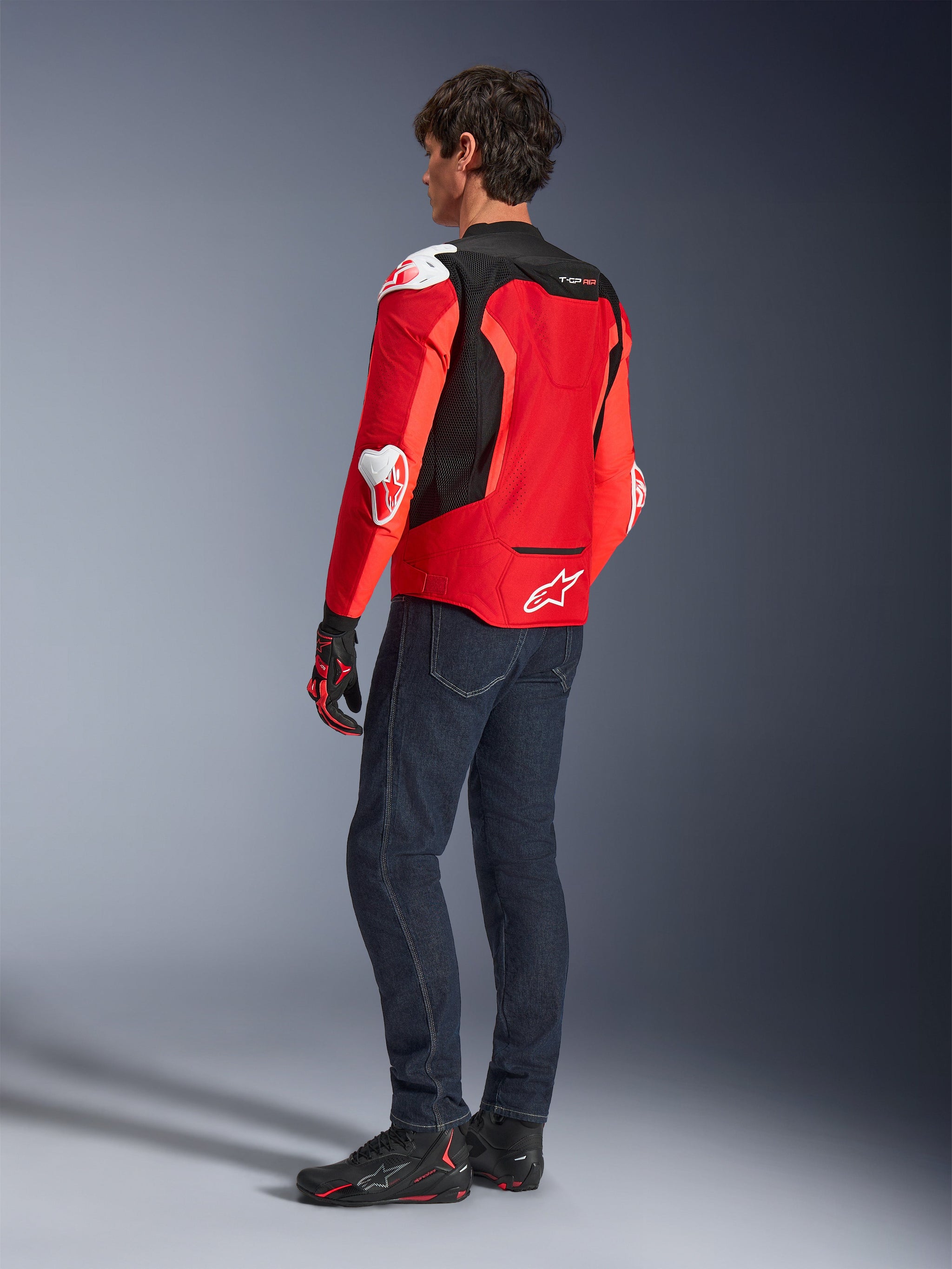 Giacca Alpinestars T-GP Air, vista posteriore di un uomo che indossa una giacca in tessuto rosso brillante e rosso fluo con pannelli in mesh nero e slider bianchi, abbinata a jeans blu scuro e scarpe da moto nere, abbigliamento tecnico da moto sportiva.