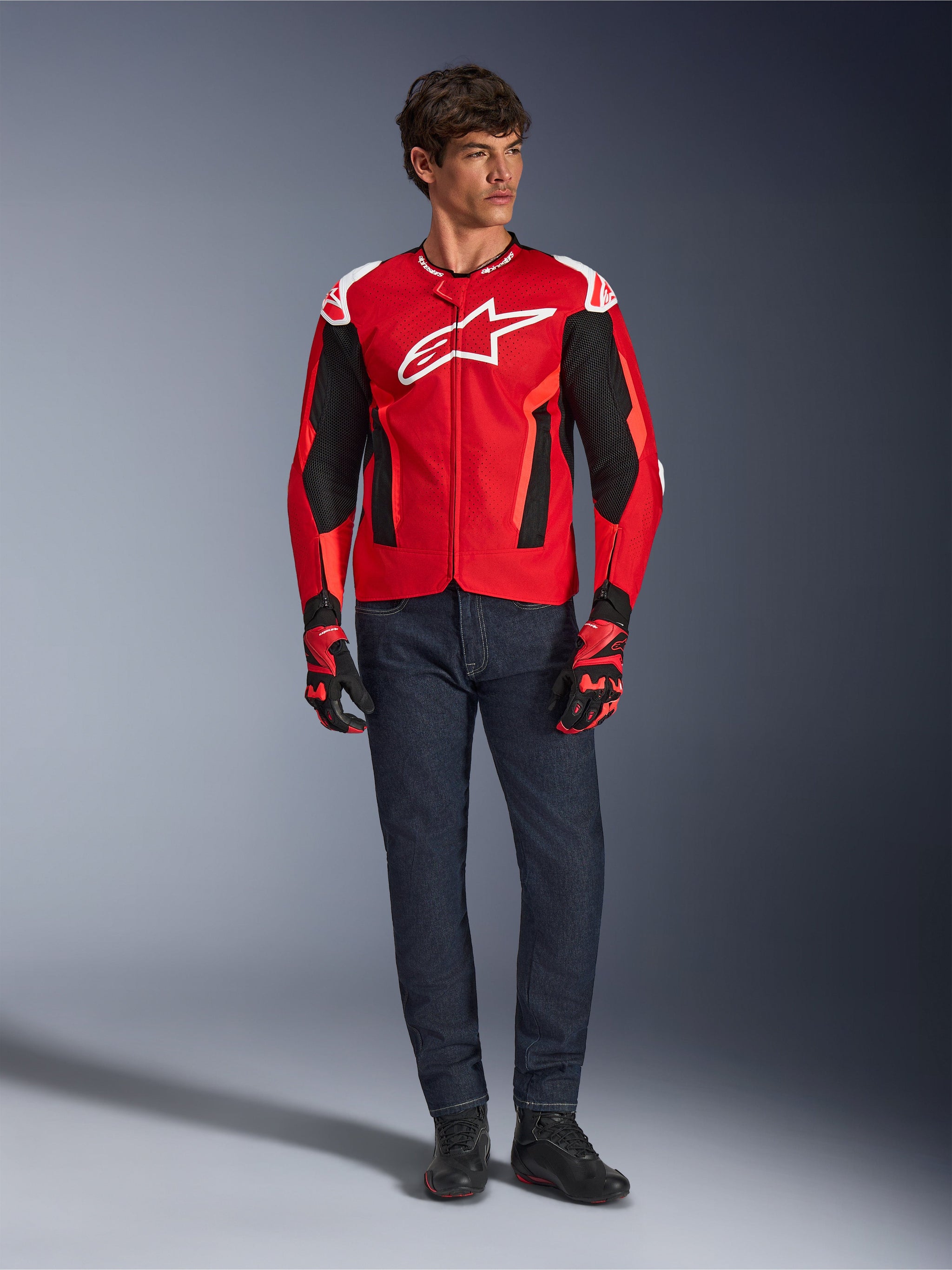 Giacca Alpinestars T-GP Air, Bright Red Red Fluo Black, modello maschile che indossa un outfit da moto completo con jeans blu scuro e guanti coordinati, giacca sportiva in tessuto con pannelli in rete traforata e logo Astars bianco, scatto in studio.
