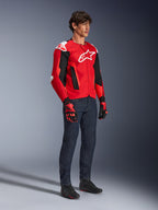 Giacca Alpinestars T-GP Air, giacca da moto sportiva, rosso acceso e nero con accenti bianchi, indossata da un modello con jeans blu scuro e guanti da corsa rossi, dotata di pannelli in mesh ad alta ventilazione e slider sulle spalle bianchi, vestibilità sportiva aerodinamica per la guida in climi caldi.