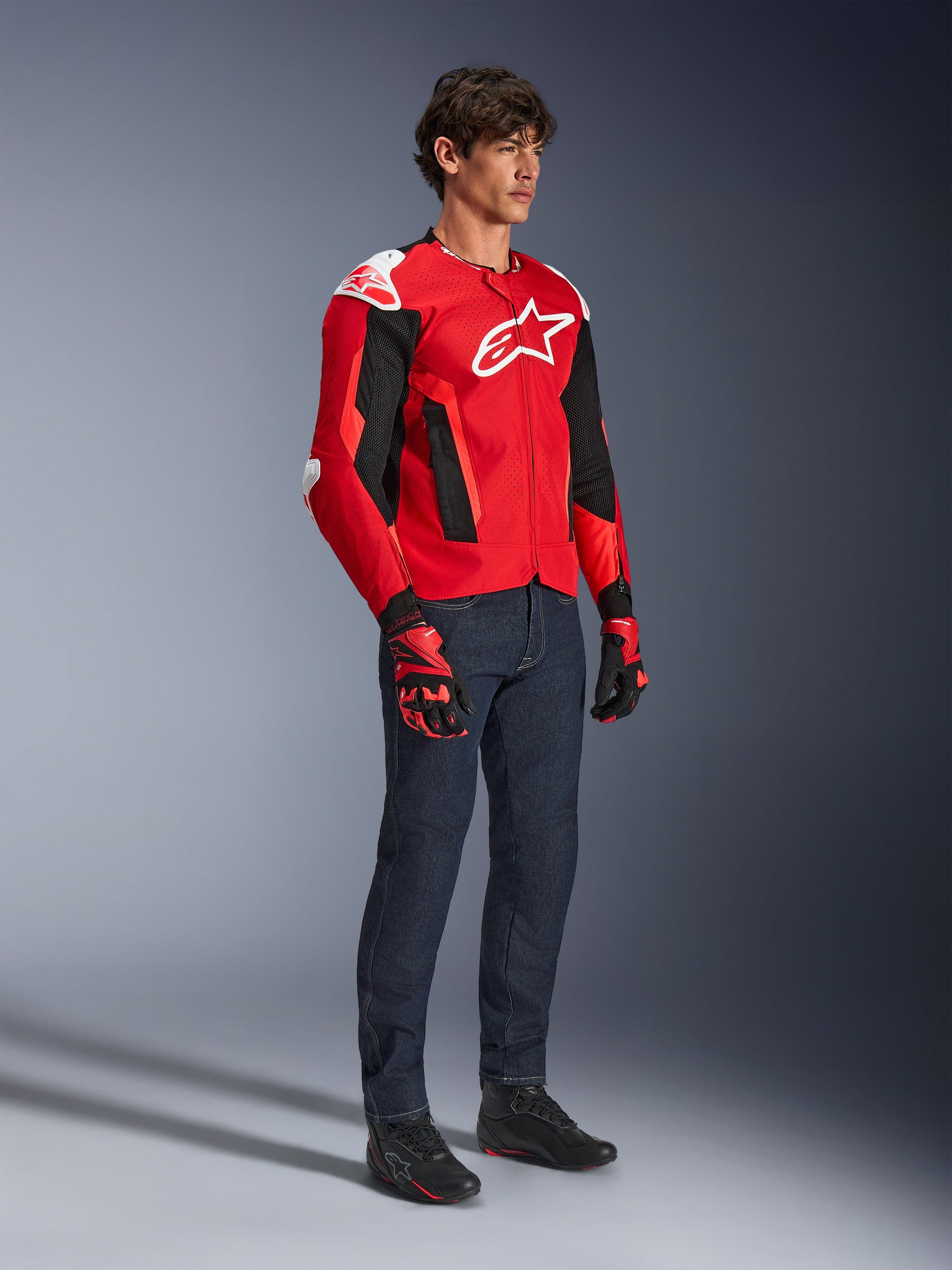 Giacca Alpinestars T-GP Air, giacca da moto sportiva, rosso acceso e nero con accenti bianchi, indossata da un modello con jeans blu scuro e guanti da corsa rossi, dotata di pannelli in mesh ad alta ventilazione e slider sulle spalle bianchi, vestibilità sportiva aerodinamica per la guida in climi caldi.