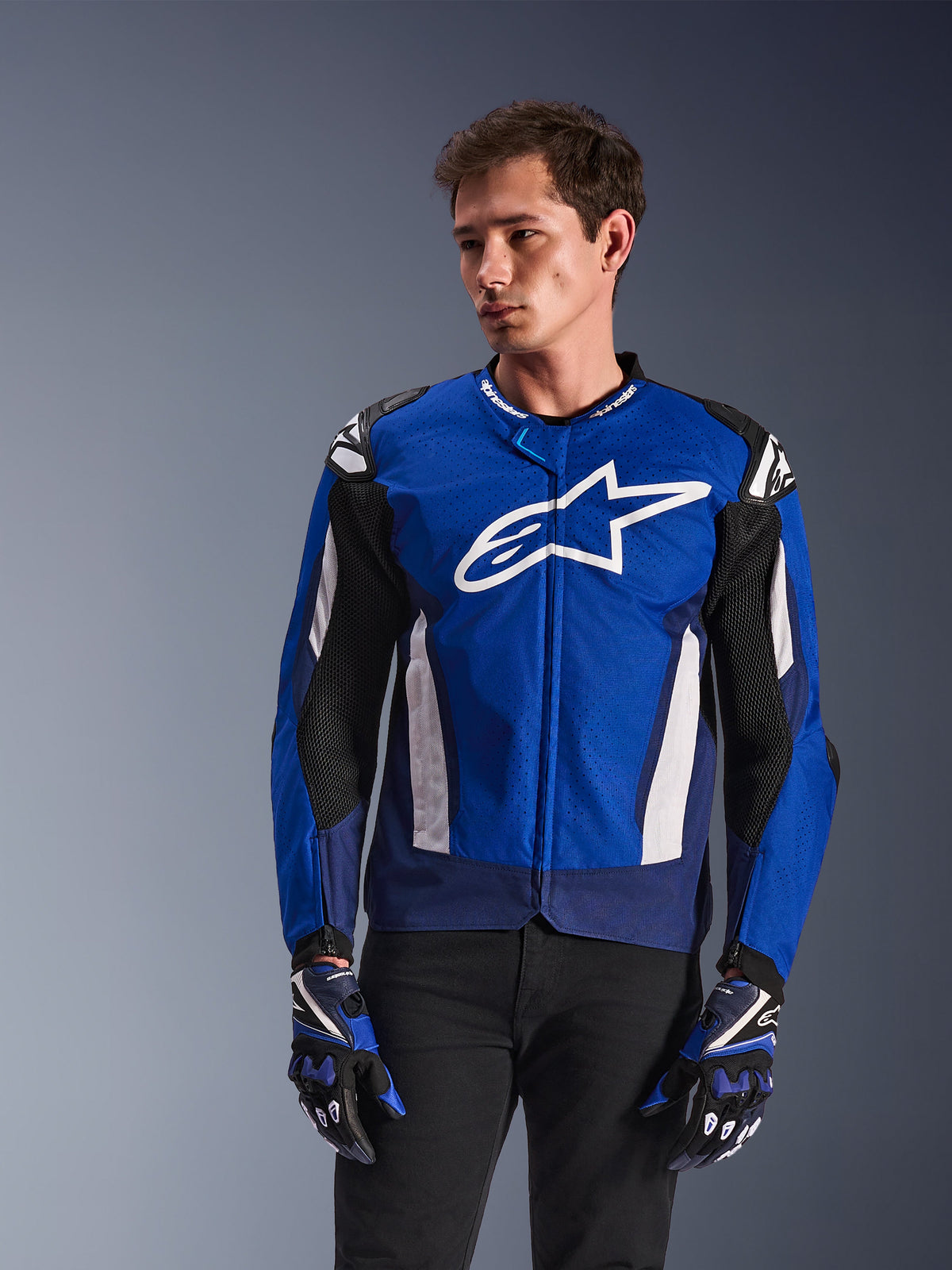 Alpinestars T-GP Air Jacket, giacca da moto sportiva in tessuto, blu brillante e navy con accenti bianchi, con grande logo a stella sul petto e pannelli di ventilazione in mesh, indossata da un modello con guanti coordinati e pantaloni neri