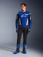 Modello che indossa la giacca Alpinestars T-GP Air in Bright Blue Navy White, abbinata a pantaloni neri, guanti da corsa coordinati blu e navy e stivali da moto blu e neri, scatto in studio che mostra un outfit completo da guida sportiva.