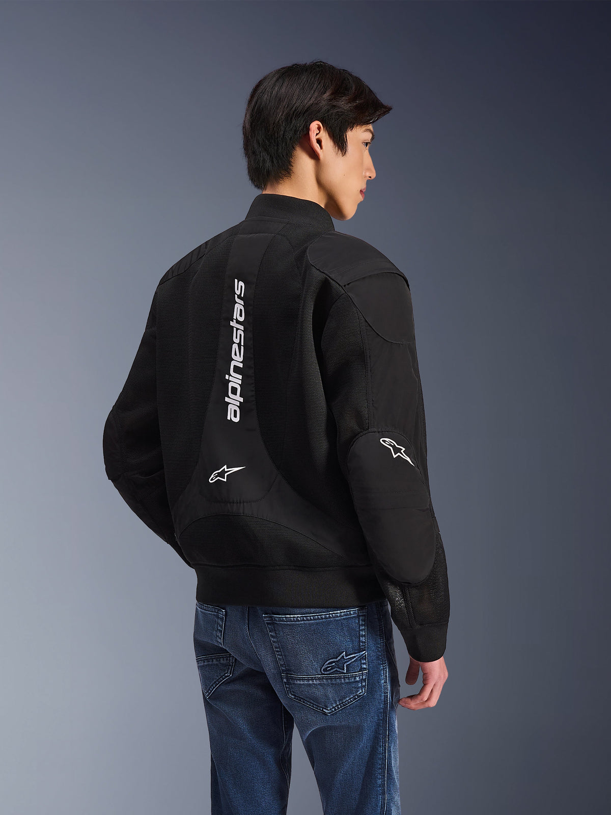 Giacca Alpinestars Flight Air, giacca da moto nera, vista posteriore con grande logo bianco verticale e pannelli di ventilazione in mesh, stile bomber con orlo a costine, indossata con jeans blu, abbigliamento tecnico da moto urbano contemporaneo