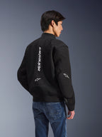 Giacca Alpinestars Flight Air, giacca da moto nera, vista posteriore con grande logo bianco verticale e pannelli di ventilazione in mesh, stile bomber con orlo a costine, indossata con jeans blu, abbigliamento tecnico da moto urbano contemporaneo