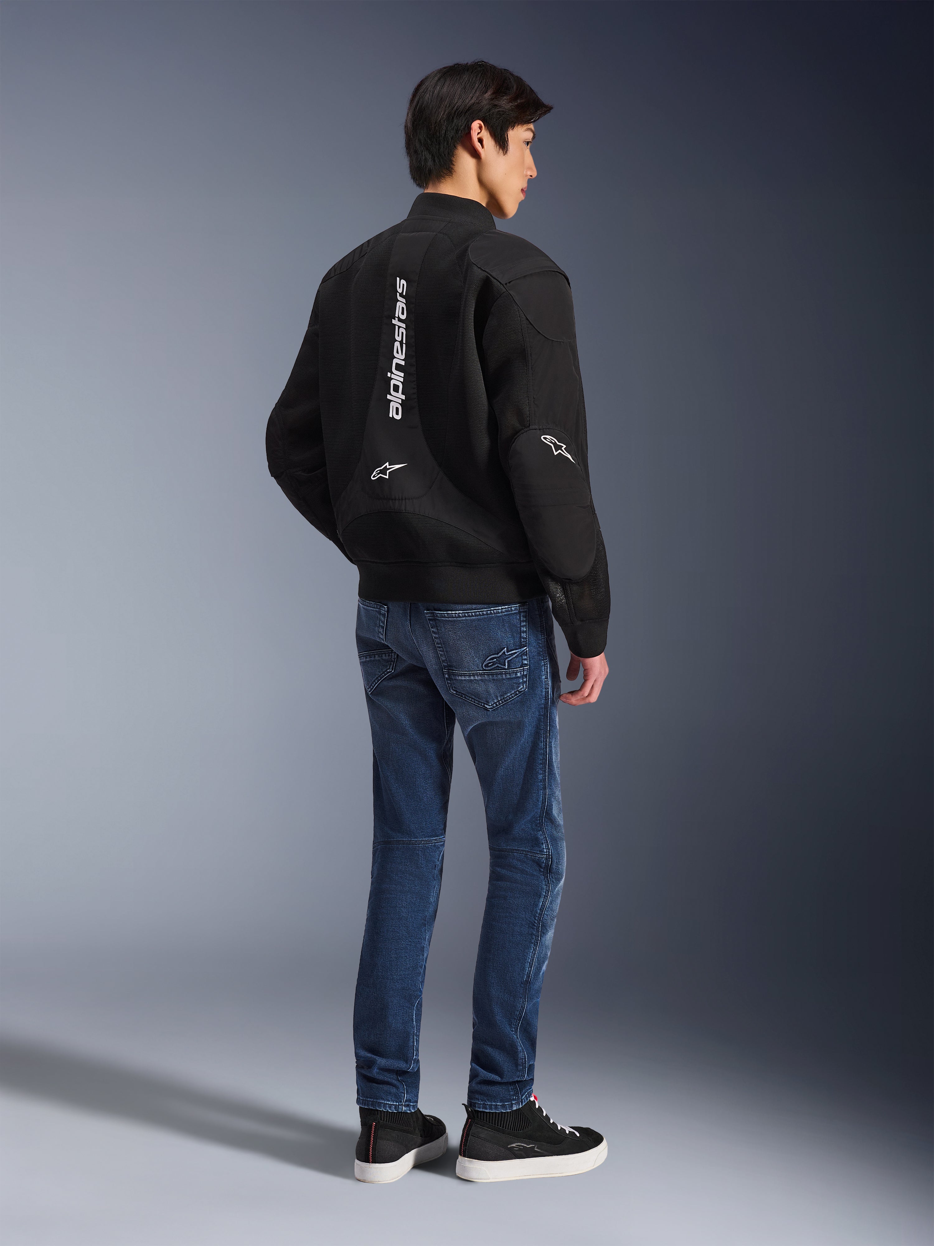 Modello che indossa la giacca Alpinestars Flight Air nera, vista dal retro, abbinata a jeans da moto blu e sneakers nere, con logo Alpinestars bianco verticale e pannelli di ventilazione in mesh 3D.