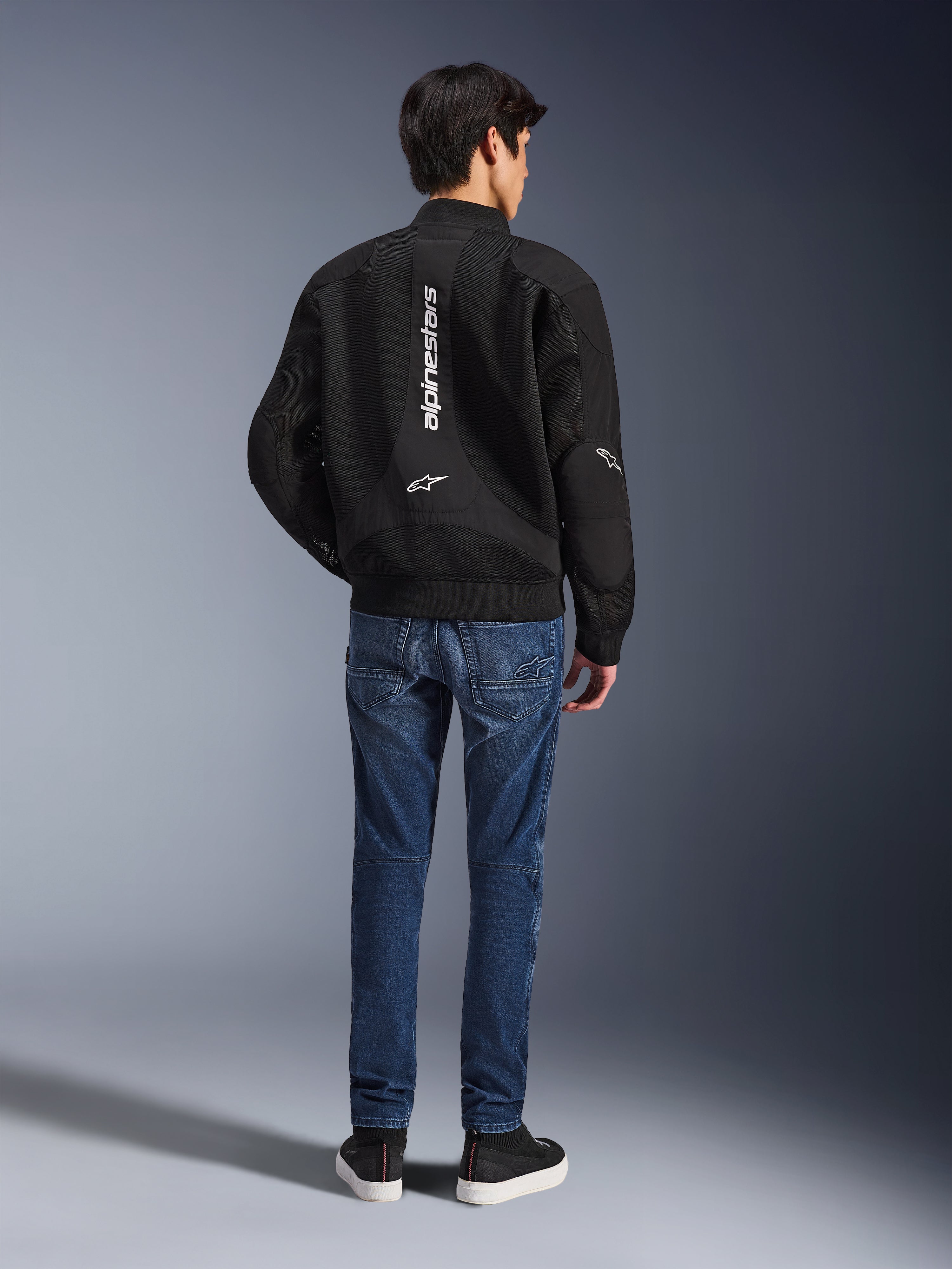 Modello che indossa la giacca Alpinestars Flight Air vista da dietro, giacca da moto nera stile bomber con pannelli in mesh 3D e logo bianco verticale, abbinata a jeans da moto in denim blu e sneakers nere, abbigliamento da moto urbano contemporaneo