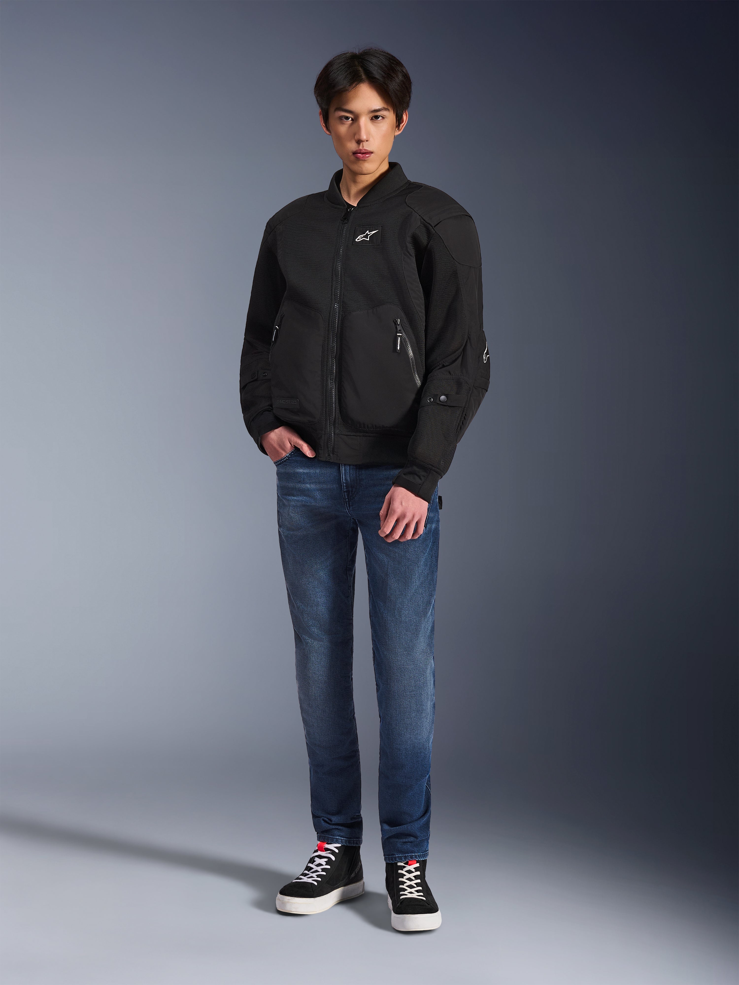 Modello che indossa la giacca Alpinestars Flight Air in nero, una giacca da moto urbana in stile bomber con pannelli in mesh 3D e cerniera frontale, abbinata a jeans blu e sneakers nere, abbigliamento da guida contemporaneo d'ispirazione street.