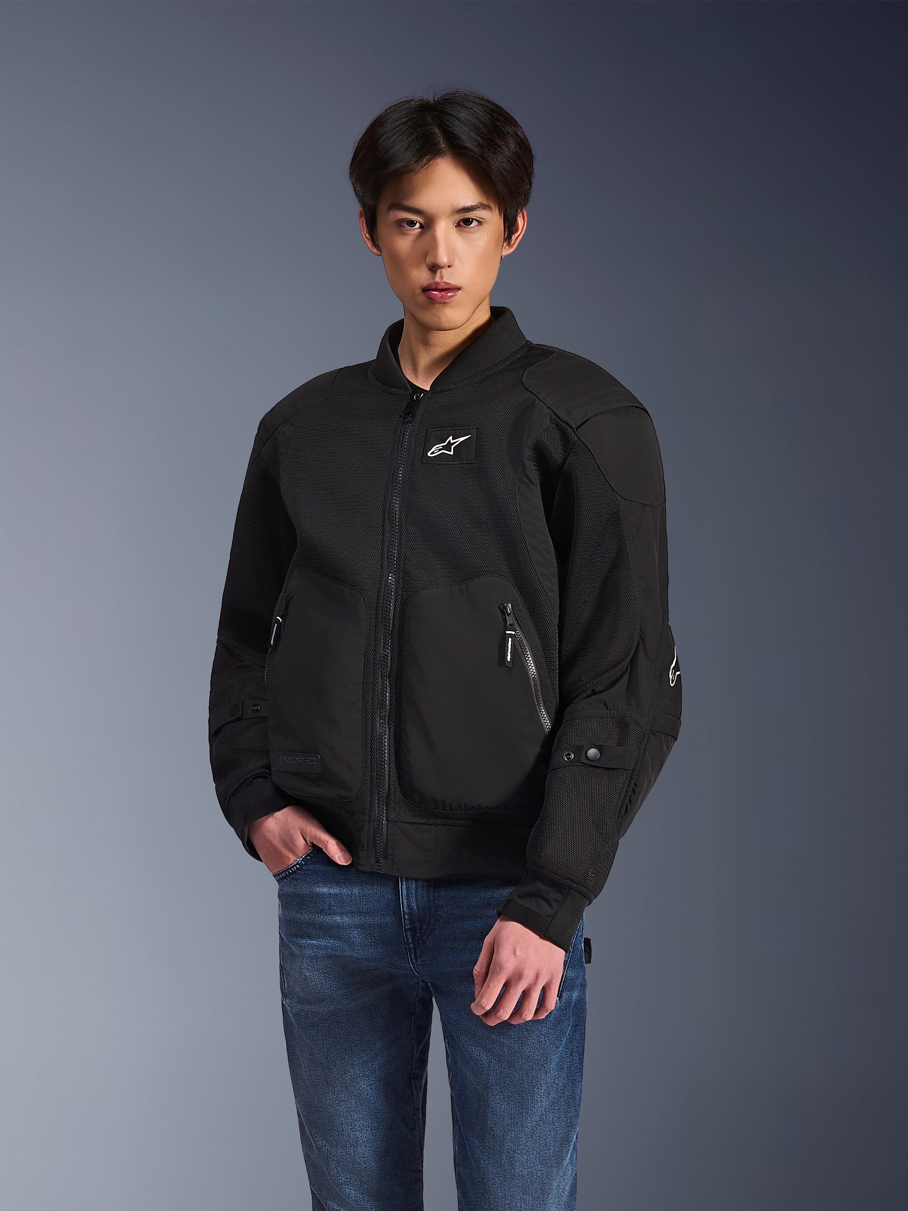 Alpinestars Flight Air Jacket, giacca da moto urbana nera, indossata da un uomo con jeans blu, caratterizzata da un'estetica stile bomber con pannelli in mesh, cerniera frontale e logo bianco sul petto, progettata per la guida urbana in climi caldi