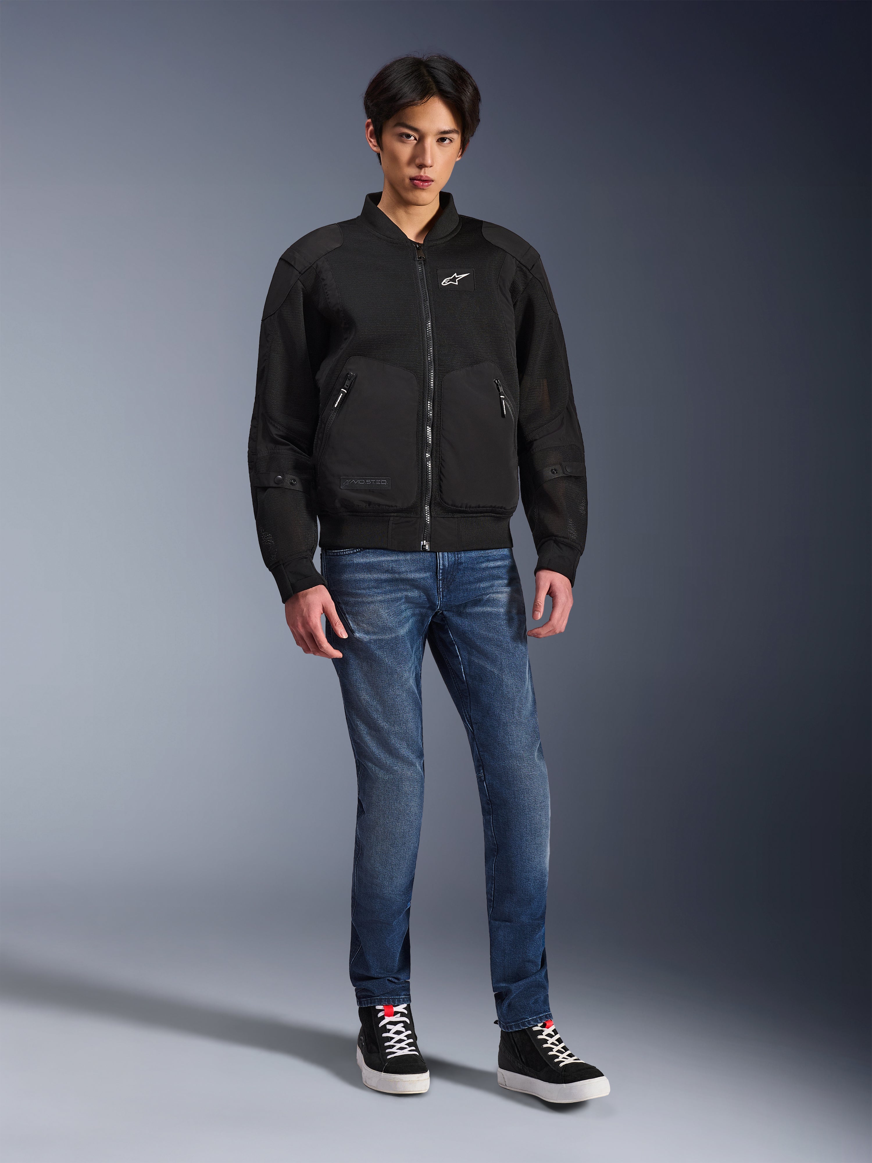 Alpinestars Flight Air Jacket, nera, giacca da moto stile bomber d'ispirazione urbana con pannelli in mesh 3D e logo bianco sul petto, indossata da un modello con jeans blu e sneakers nere, abbigliamento da moto contemporaneo