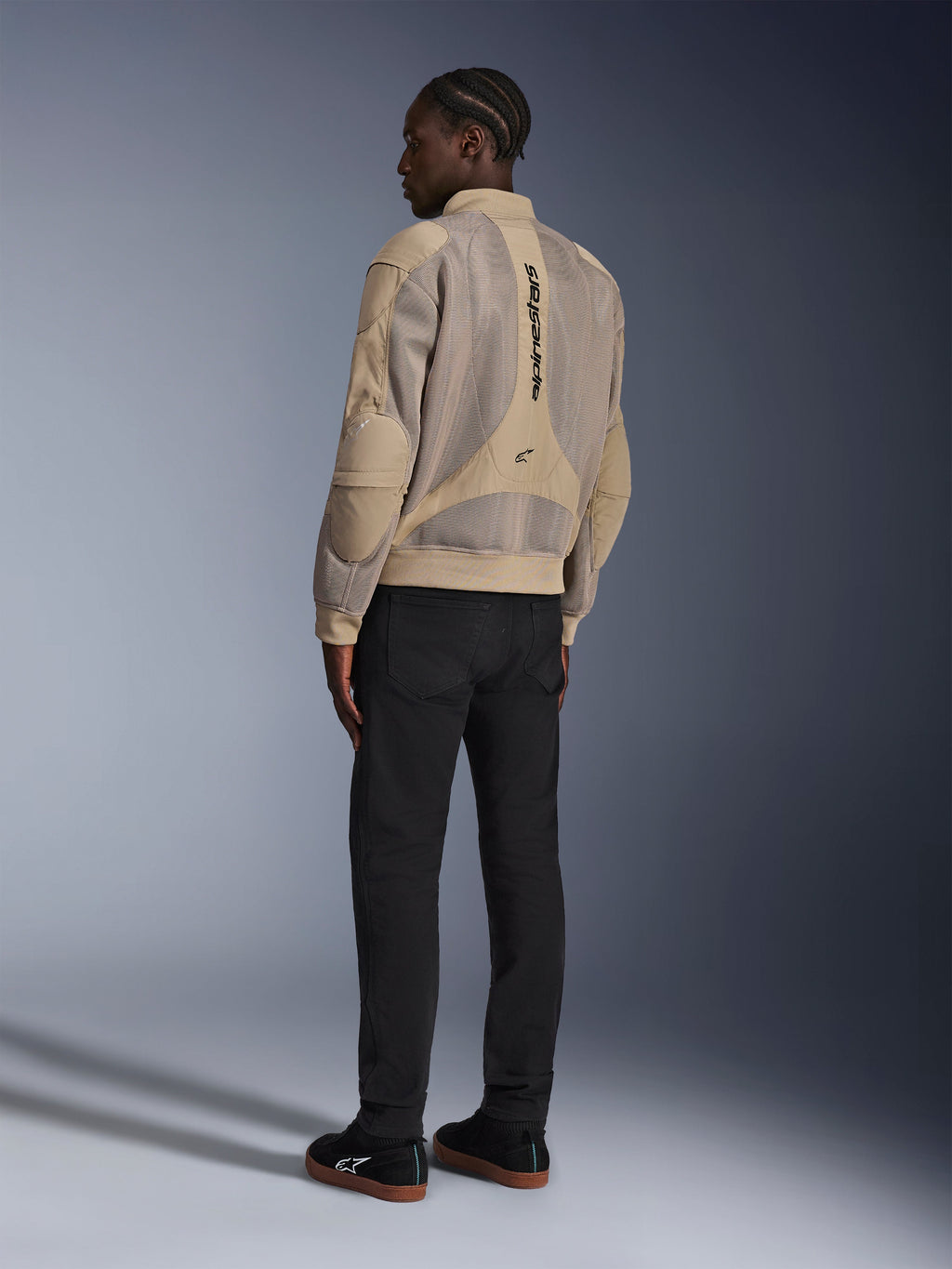 Giacca Alpinestars Flight Air, giacca da moto urbana in tessuto, marrone, vista posteriore che mostra ampi pannelli in mesh 3D e logo verticale, silhouette stile bomber con colletto e orlo a costine, abbinata a pantaloni neri e scarpe nere per uno stile di guida di ispirazione street.