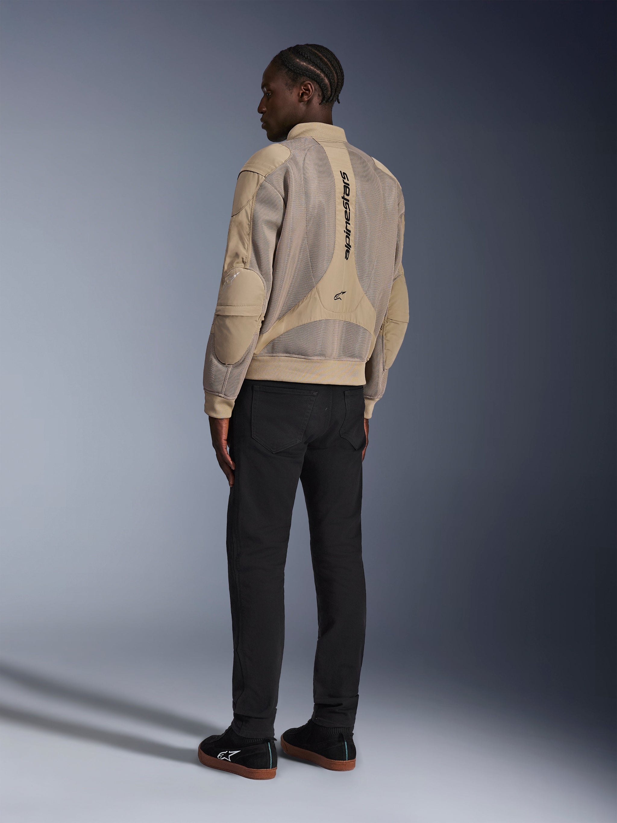 Giacca Alpinestars Flight Air, giacca da moto urbana in tessuto, marrone, vista posteriore che mostra ampi pannelli in mesh 3D e logo verticale, silhouette stile bomber con colletto e orlo a costine, abbinata a pantaloni neri e scarpe nere per uno stile di guida di ispirazione street.