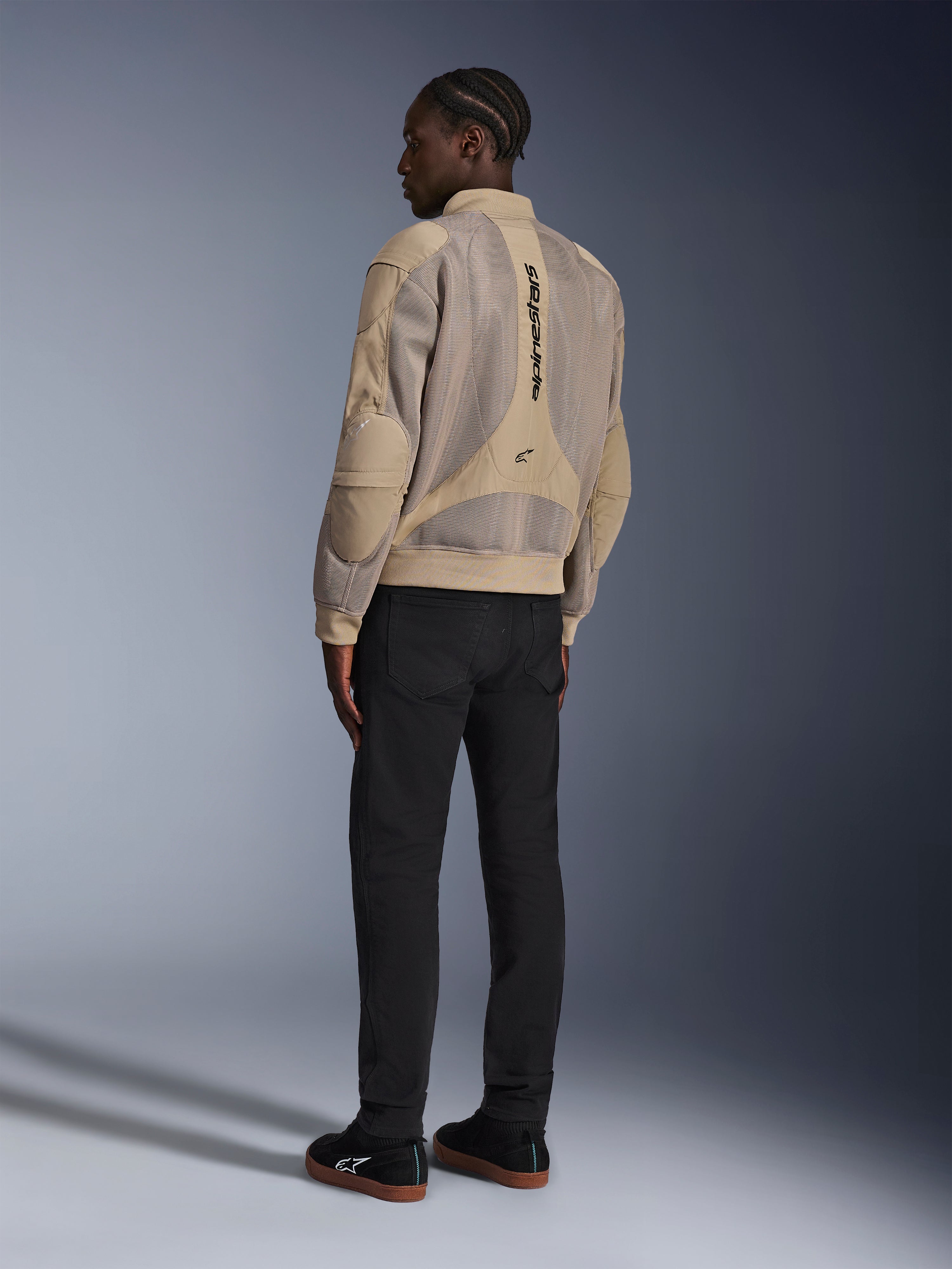 Giacca Alpinestars Flight Air, giacca da moto urbana in tessuto, marrone, vista posteriore che mostra ampi pannelli in mesh 3D e logo verticale, silhouette stile bomber con colletto e orlo a costine, abbinata a pantaloni neri e scarpe nere per uno stile di guida di ispirazione street.