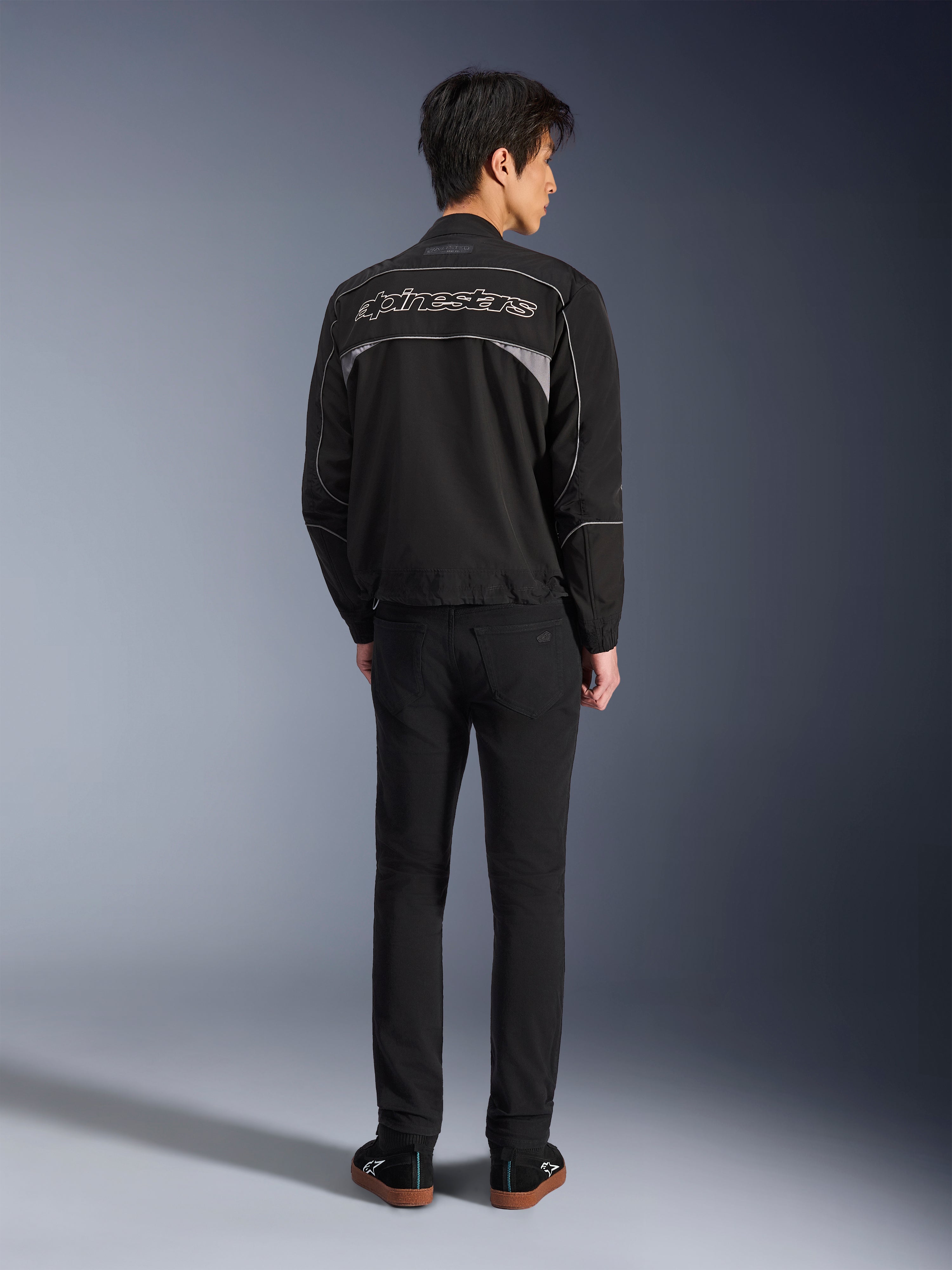 Giacca Alpinestars Unite, giacca da moto nero e grigio scuro, vista posteriore con logo Alpinestars profilato sulla schiena, pannelli riflettenti grigi, design urbano minimalista, indossata con pantaloni neri e scarpe nere