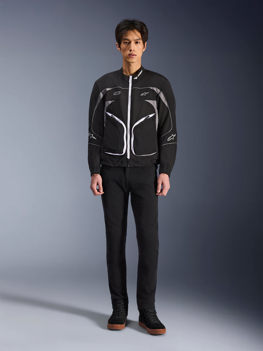Giacca Alpinestars Unite, giacca da moto, nero e grigio scuro, indossata da un modello con pantaloni neri e stivali neri, con cerniere bianche a contrasto, profili riflettenti e design urbano minimalista