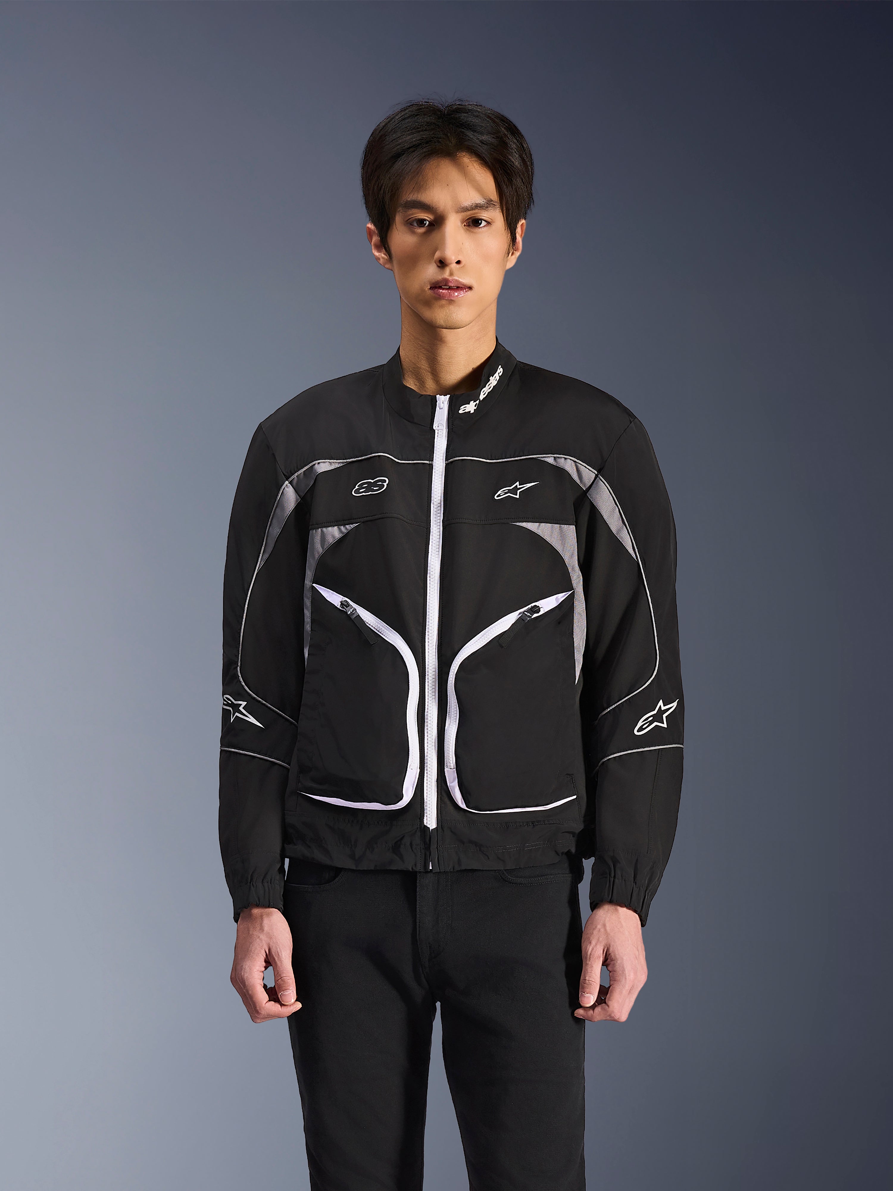 Giacca Alpinestars Unite, giacca da moto urbana minimalista, nero e grigio scuro con accenti bianchi, dotata di cerniera anteriore bianca, profili riflettenti e logo Alpinestars su petto e maniche, stile streetwear contemporaneo per motociclisti
