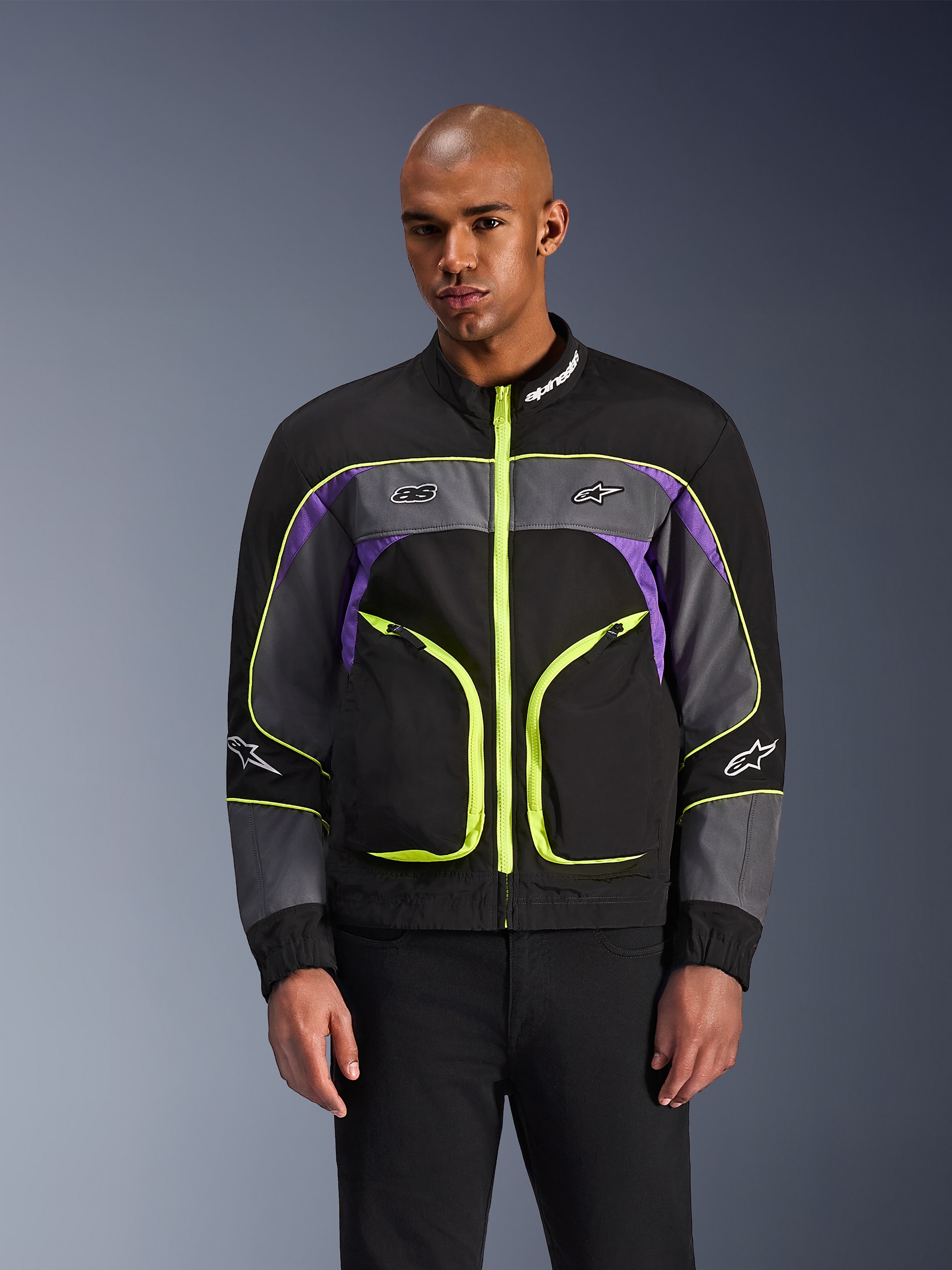 Alpinestars Unite Jacket, giacca da moto urbana, Nero Asphalt e Viola con accenti giallo neon, ampie tasche anteriori con zip neon, colletto alto con logo bianco, indossata da un modello, abbigliamento protettivo contemporaneo ispirato allo streetwear