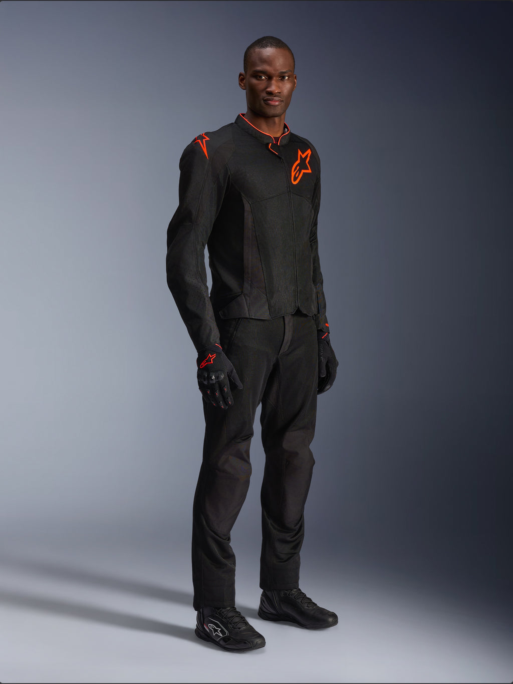 Modello che indossa la giacca Alpinestars Viper Air V4 in nero e rosso fluo, con pannelli in rete e loghi a stella rossi sul petto e sulla spalla, abbinata a pantaloni da moto, guanti e scarpe da guida neri coordinati, completo professionale per la guida su strada