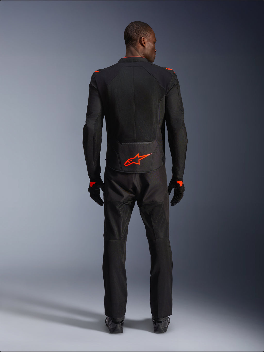 Alpinestars Viper Air V4 Jacket, nero e rosso fluo, vista posteriore su modello, indossata con pantaloni da moto e guanti neri coordinati, caratterizzata da un vivace logo rosso fluorescente sulla parte bassa della schiena, costruzione in rete per il flusso d'aria, vestibilità atletica per la guida su strada in moto.