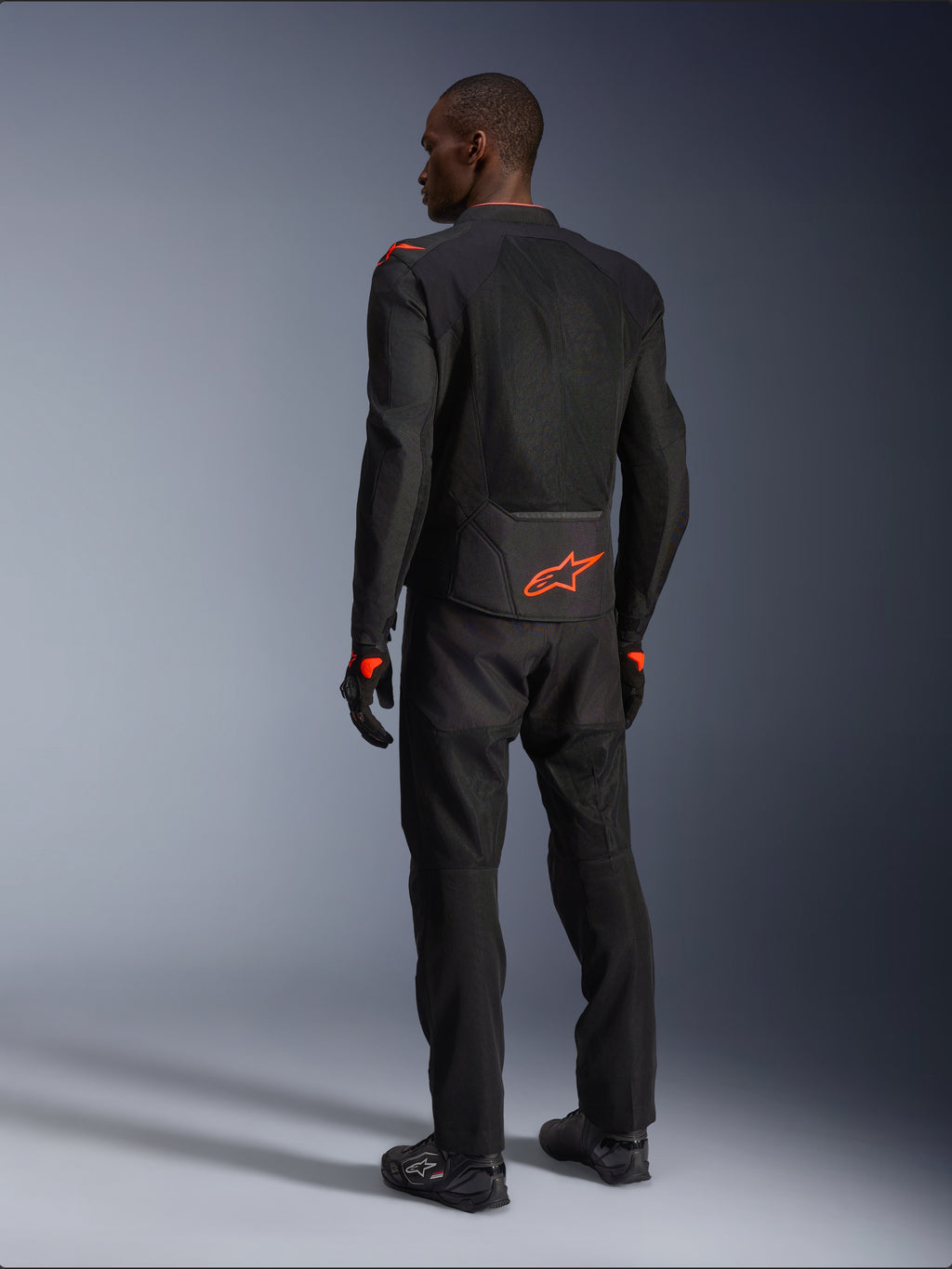 Pilota uomo visto da dietro che indossa la giacca Alpinestars Viper Air V4, nero e rosso fluo, con logo Astars arancione sulla parte bassa della schiena e sulle spalle, costruzione in tessuto mesh, abbinata a pantaloni e stivali da moto neri coordinati.