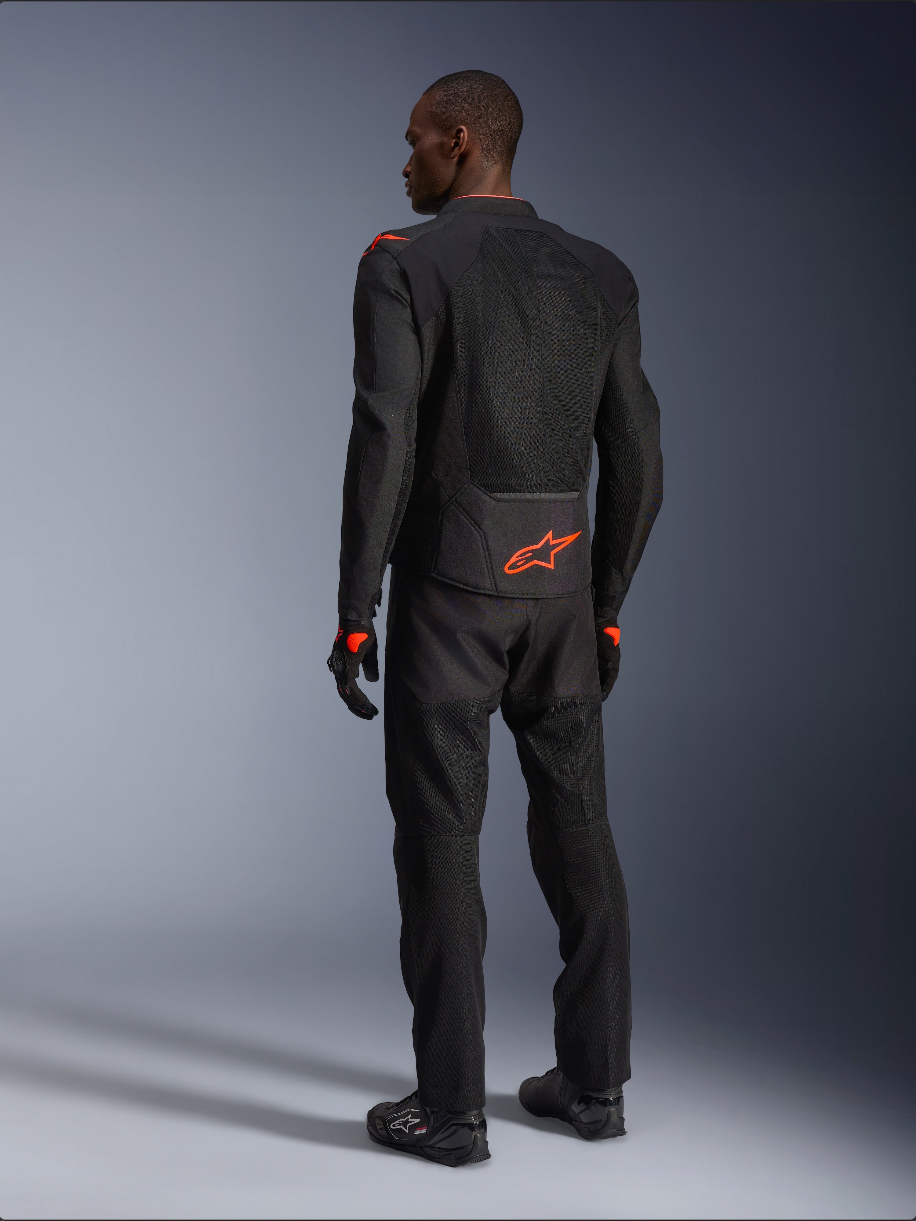 Pilota uomo visto da dietro che indossa la giacca Alpinestars Viper Air V4, nero e rosso fluo, con logo Astars arancione sulla parte bassa della schiena e sulle spalle, costruzione in tessuto mesh, abbinata a pantaloni e stivali da moto neri coordinati.