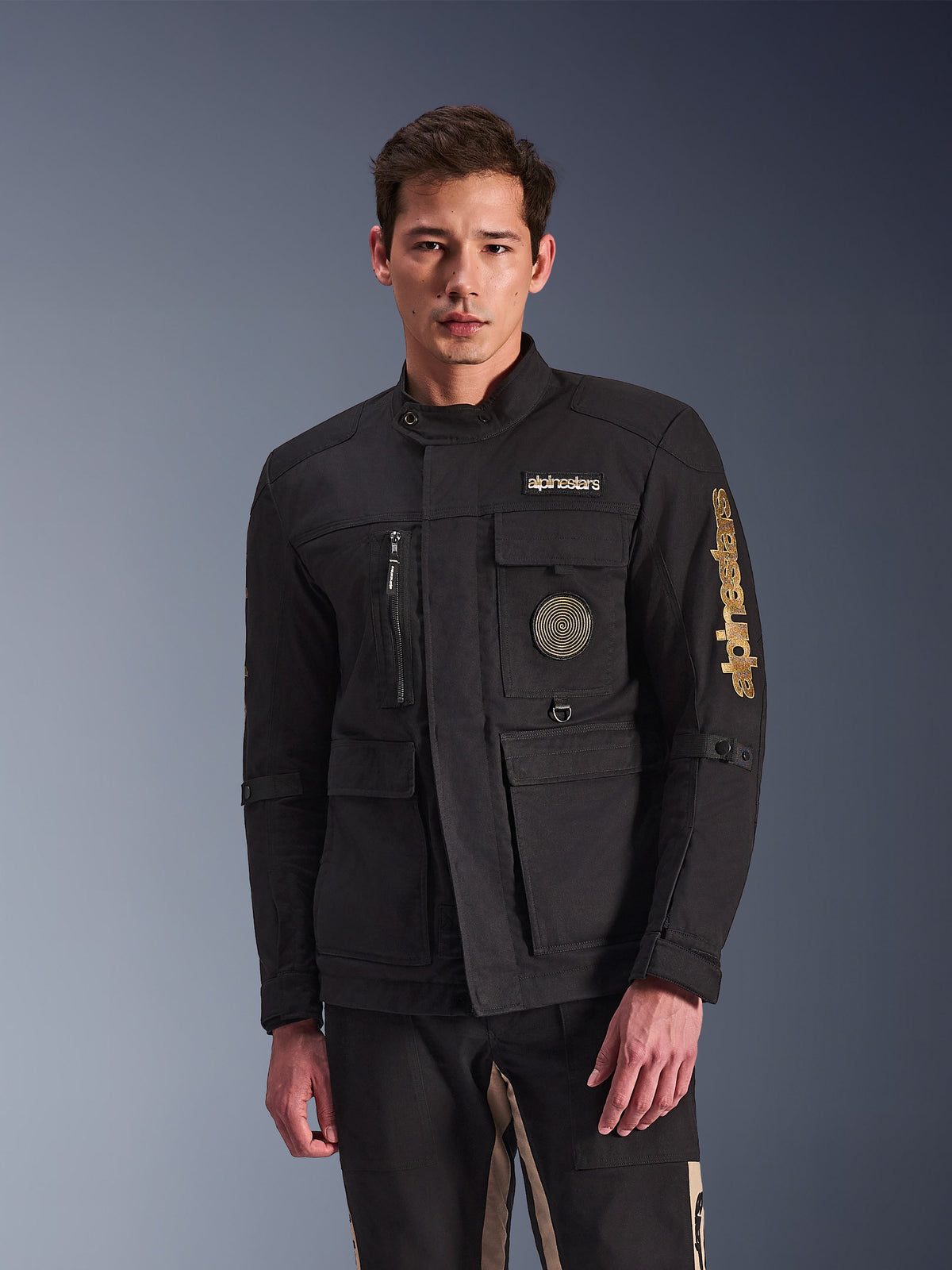 Alpinestars Dice Jacket, giacca da moto nera indossata da un modello, con loghi Alpinestars dorati, tasche funzionali multiple, patch circolare a spirale e stile urban field jacket