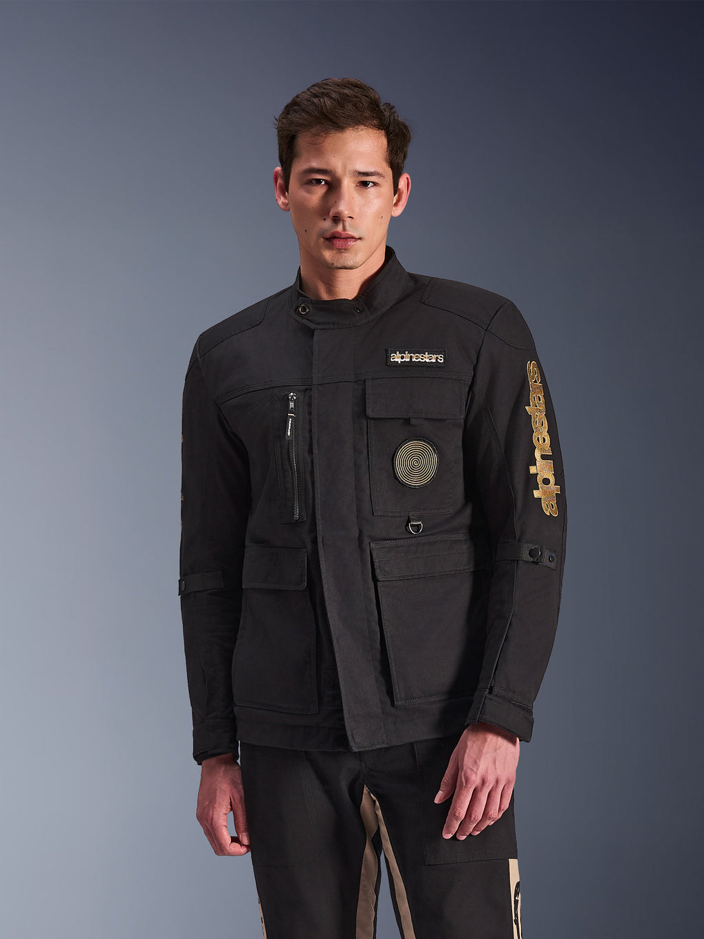 Alpinestars Dice Jacket, giacca da moto nera indossata da un modello, con loghi Alpinestars dorati, tasche funzionali multiple, patch circolare a spirale e stile urban field jacket