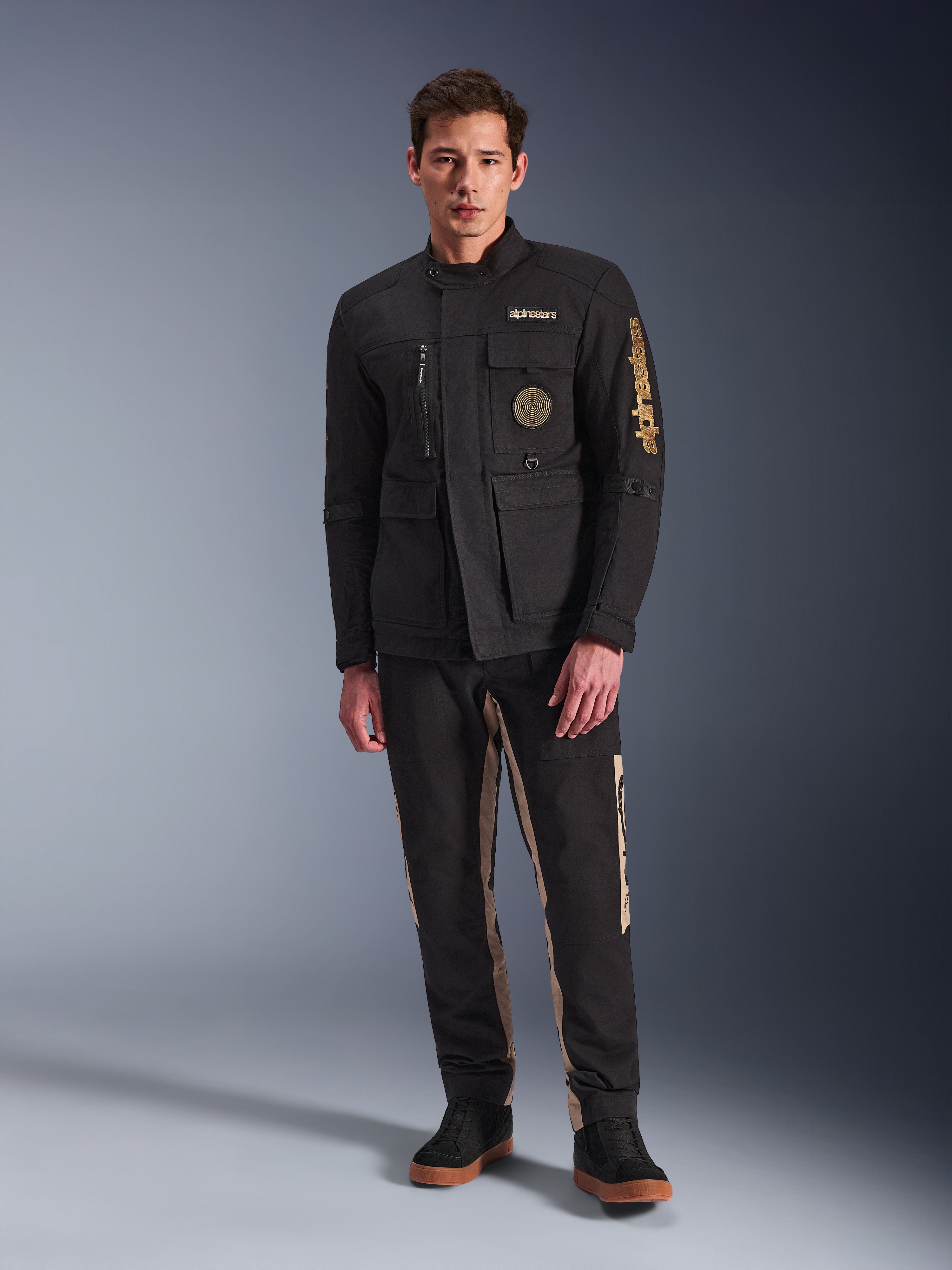 Modello che indossa la giacca Alpinestars Dice Jacket, giacca da moto urbana nera con stile vintage da field jacket, quattro tasche anteriori funzionali, logo Alpinestars sulla manica e sul petto, abbinata a pantaloni neri e beige, abbigliamento moderno per la guida urbana