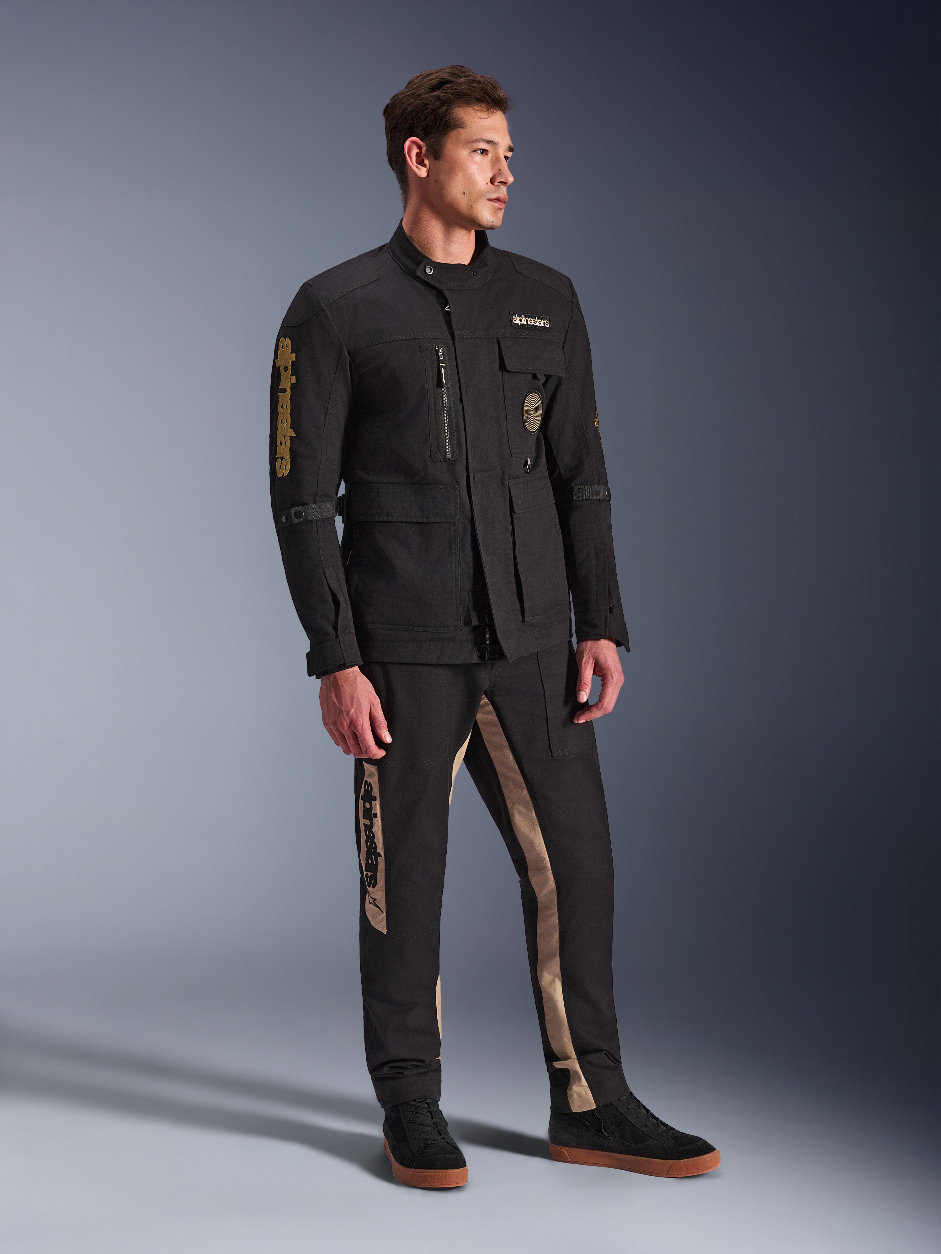 Modello che indossa la giacca Alpinestars Dice Jacket in nero, giacca da moto urbana dall'estetica vintage field jacket, con tasche utility multiple e logo dorato sulla manica, abbinata a pantaloni coordinati neri e tan e sneaker nere.