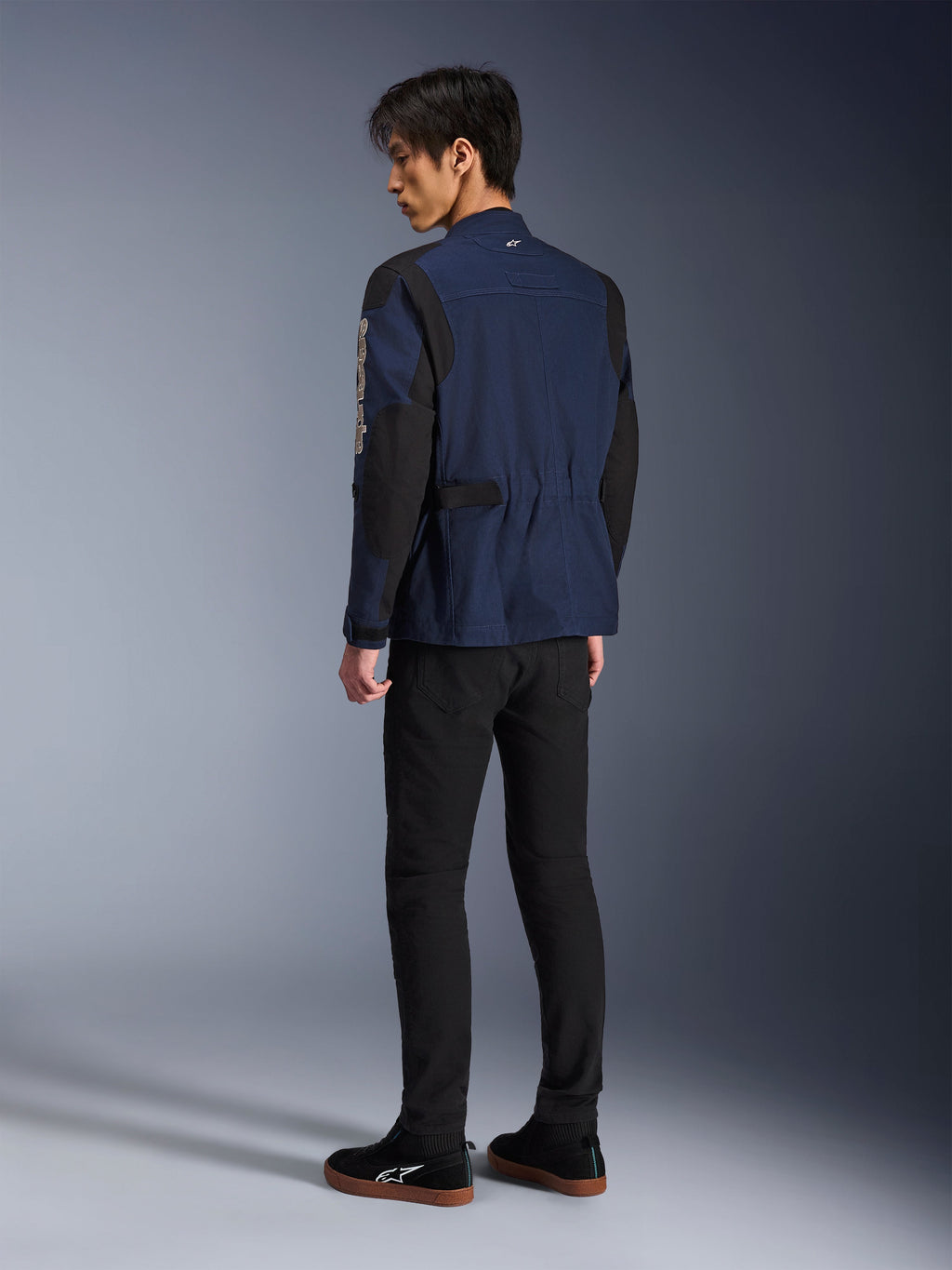 Modello uomo visto di spalle che indossa la giacca Alpinestars Dice in blu scuro e nero, abbinata a pantaloni neri e sneaker nere, giacca da moto urbana stile field jacket con spalle plissettate e linguette di regolazione in vita.