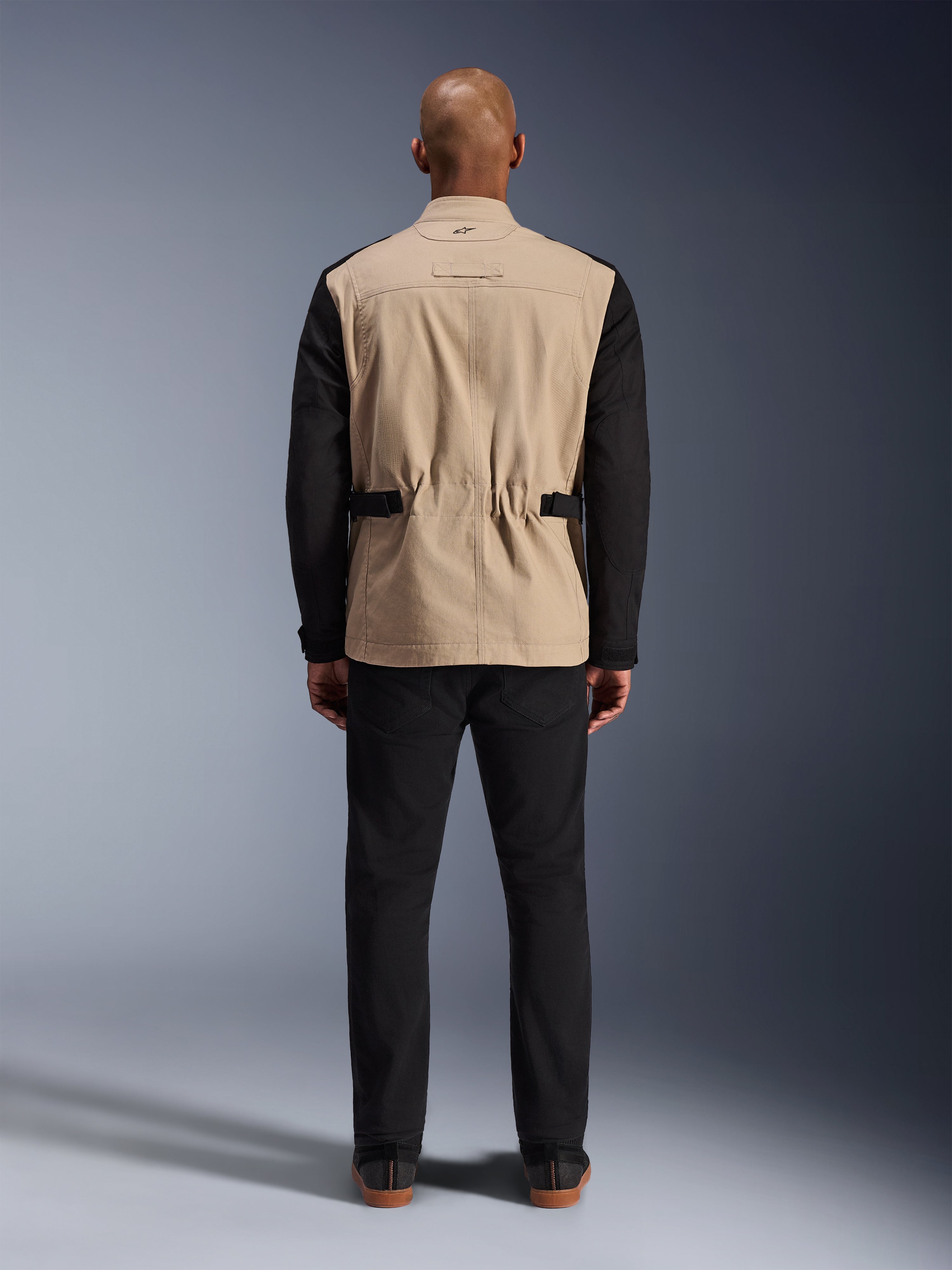 Giacca Alpinestars Dice, giacca da moto urbana, color sabbia e nero, vista posteriore su modello, caratterizzata da una struttura in twill di policotone, cinturini regolabili in vita e piccolo logo ricamato sul colletto, indossata con pantaloni neri