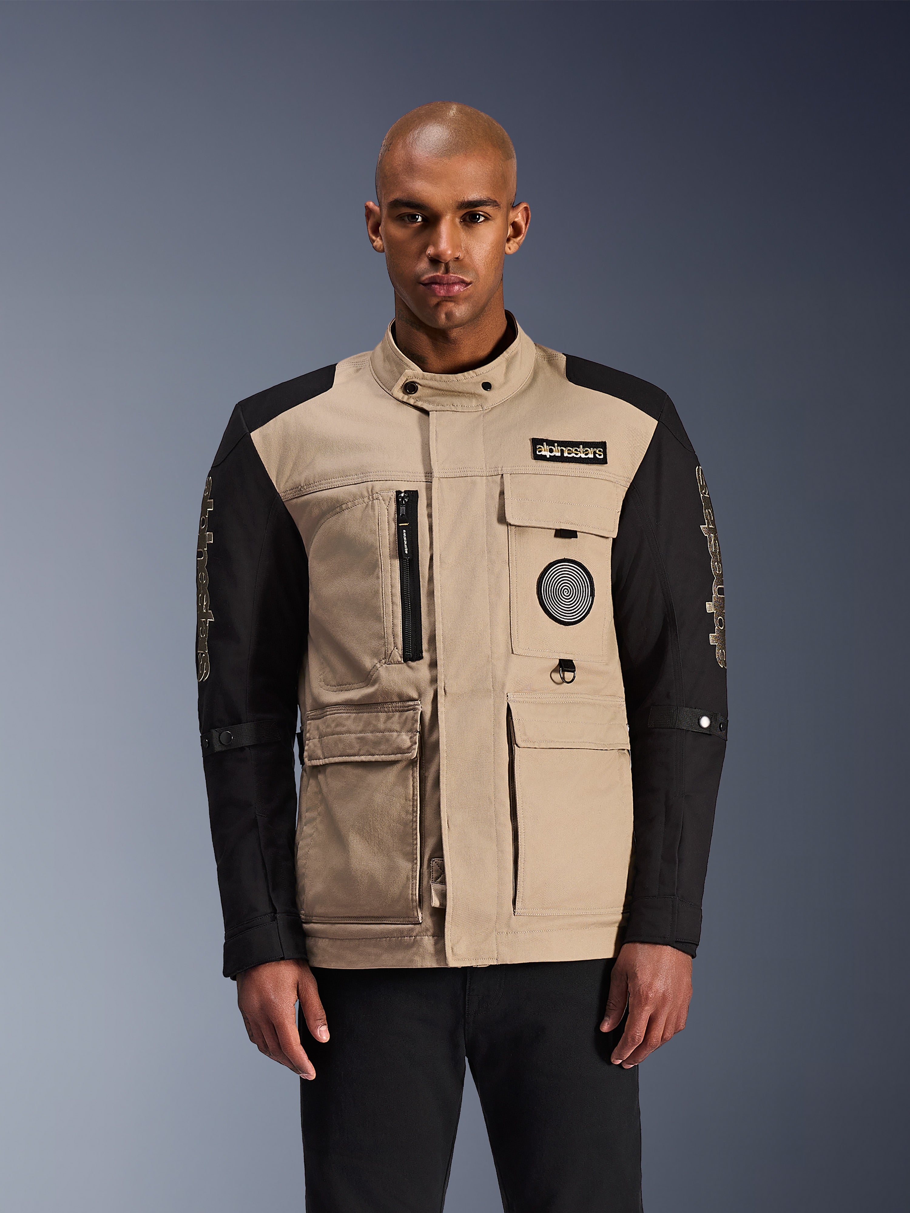 Alpinestars Dice Jacket, giacca moto urbana, beige e nero, estetica workwear robusta con tasche utility multiple e patch sul petto, colletto alto con chiusura a bottone, branding vistoso sulle maniche, indossata da un modello