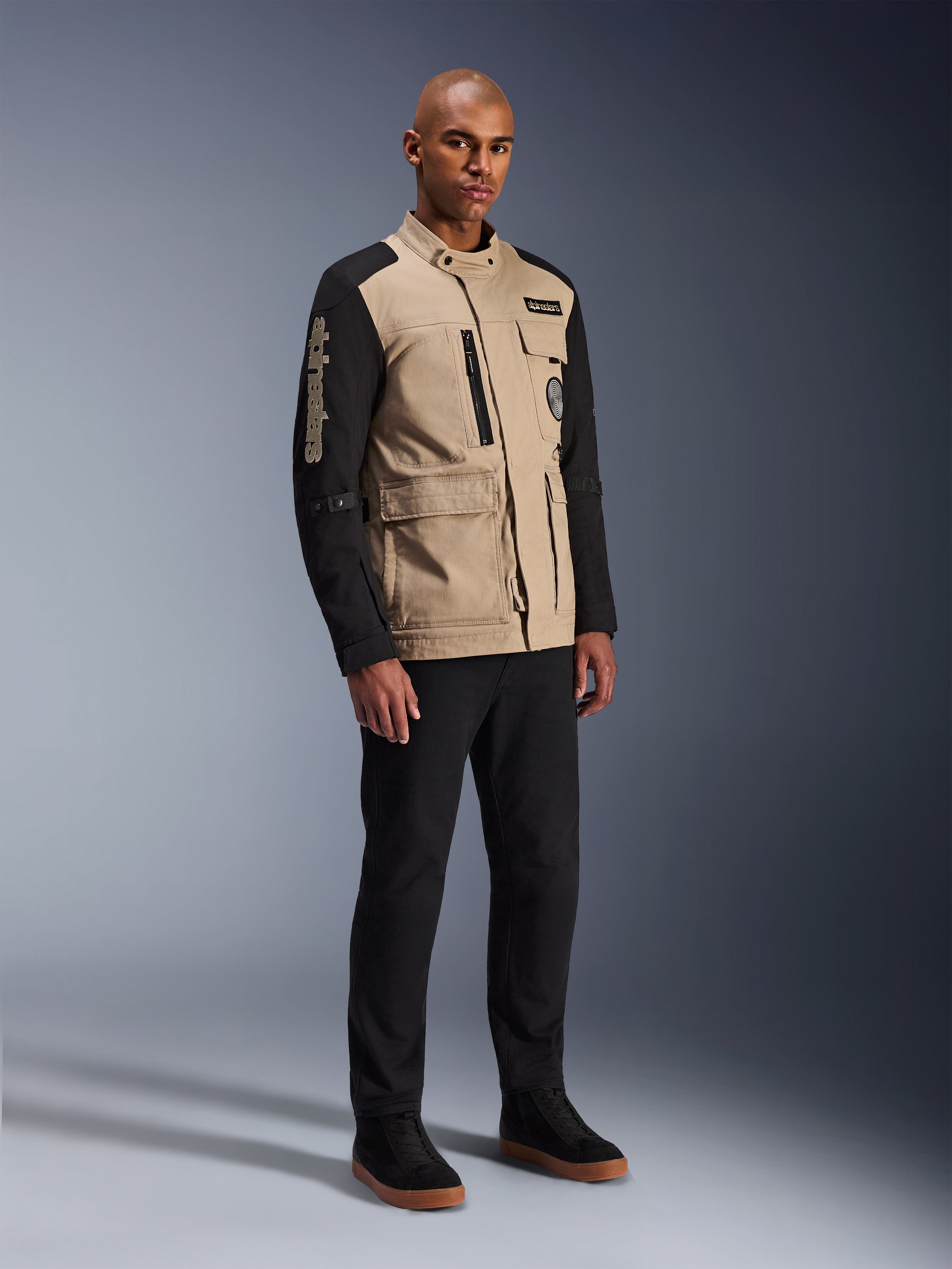 Alpinestars Giacca Dice, giacca da moto urbana, Aluminum Black e tan, design bicolore con maniche nere e busto khaki, indossata da un modello con pantaloni neri e sneakers nere, estetica ispirata al workwear con tasche utility e marchio ricamato sulla manica.
