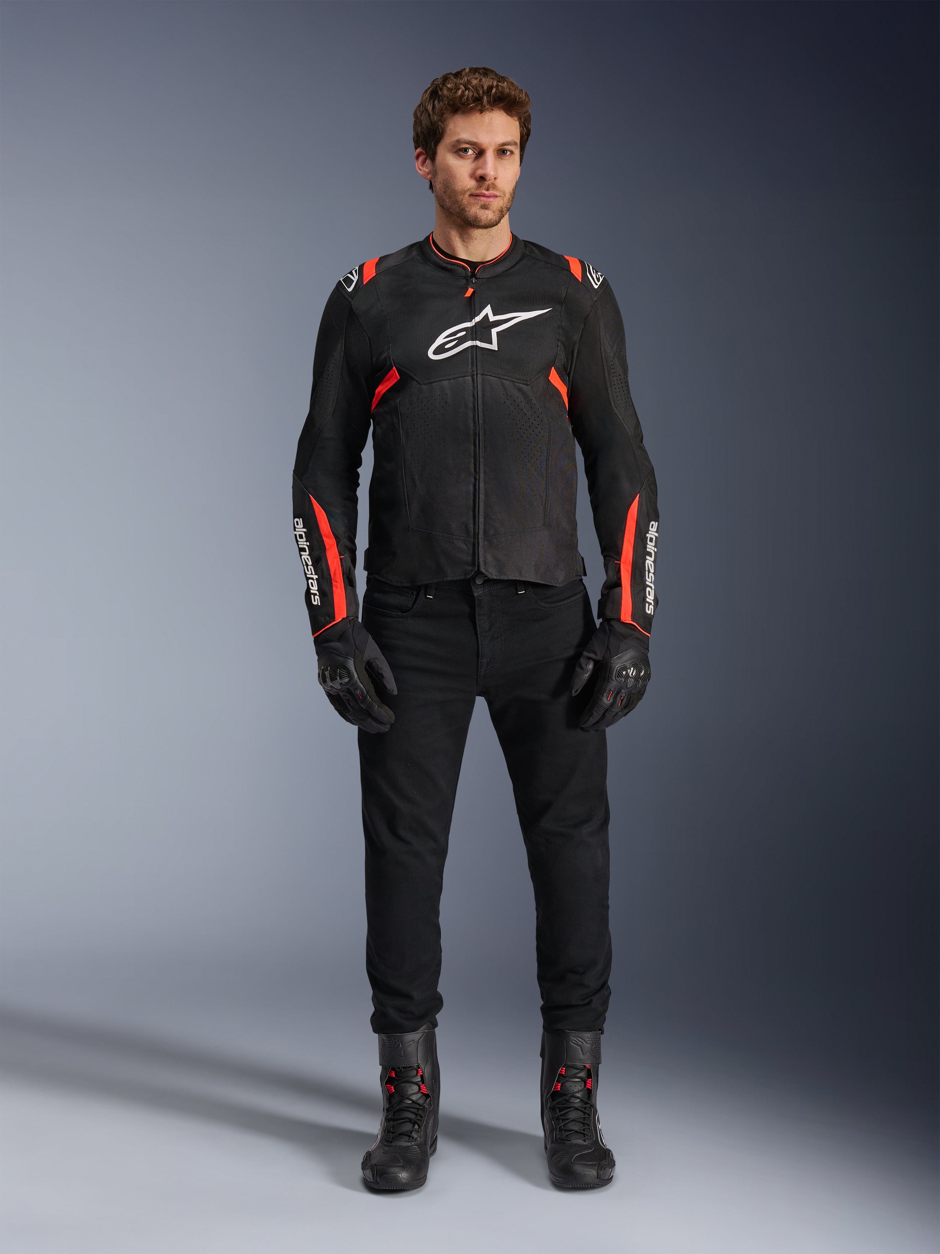 Modello che indossa la giacca Alpinestars T-SPS Air v2, giacca da moto in tessuto nero con dettagli rosso fluo, logo bianco sul petto, abbinata a pantaloni, guanti e stivali neri, stile di guida urban sport