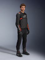 Modello che indossa la giacca Alpinestars T-Sps Air V2, nera con accenti rosso fluo, abbinata a pantaloni da moto neri, guanti e stivali neri, mettendo in risalto il design sportivo con pannelli in rete e loghi Alpinestars bianchi.