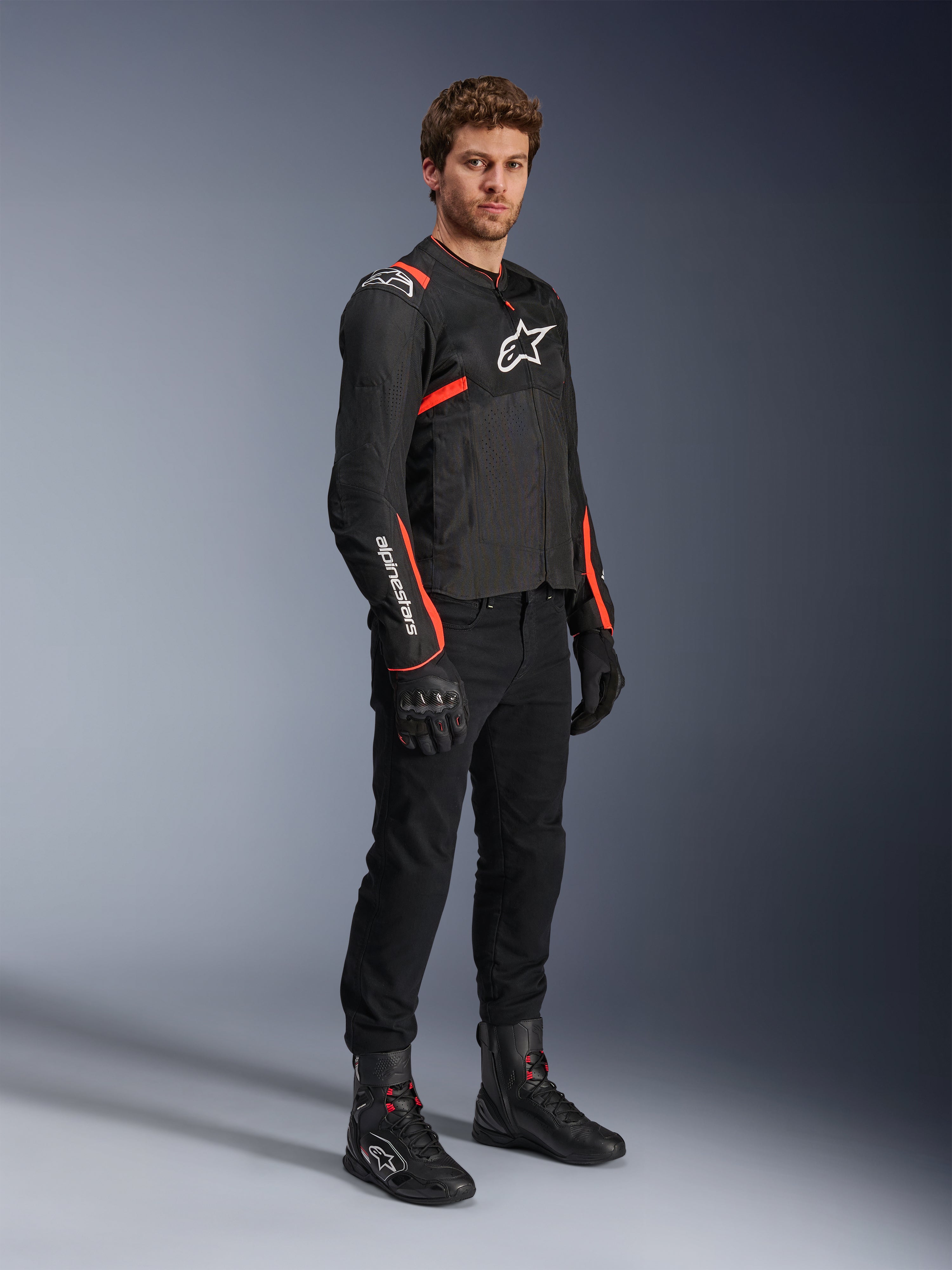 Modello che indossa la giacca Alpinestars T-Sps Air V2, nera con accenti rosso fluo, abbinata a pantaloni da moto neri, guanti e stivali neri, mettendo in risalto il design sportivo con pannelli in rete e loghi Alpinestars bianchi.