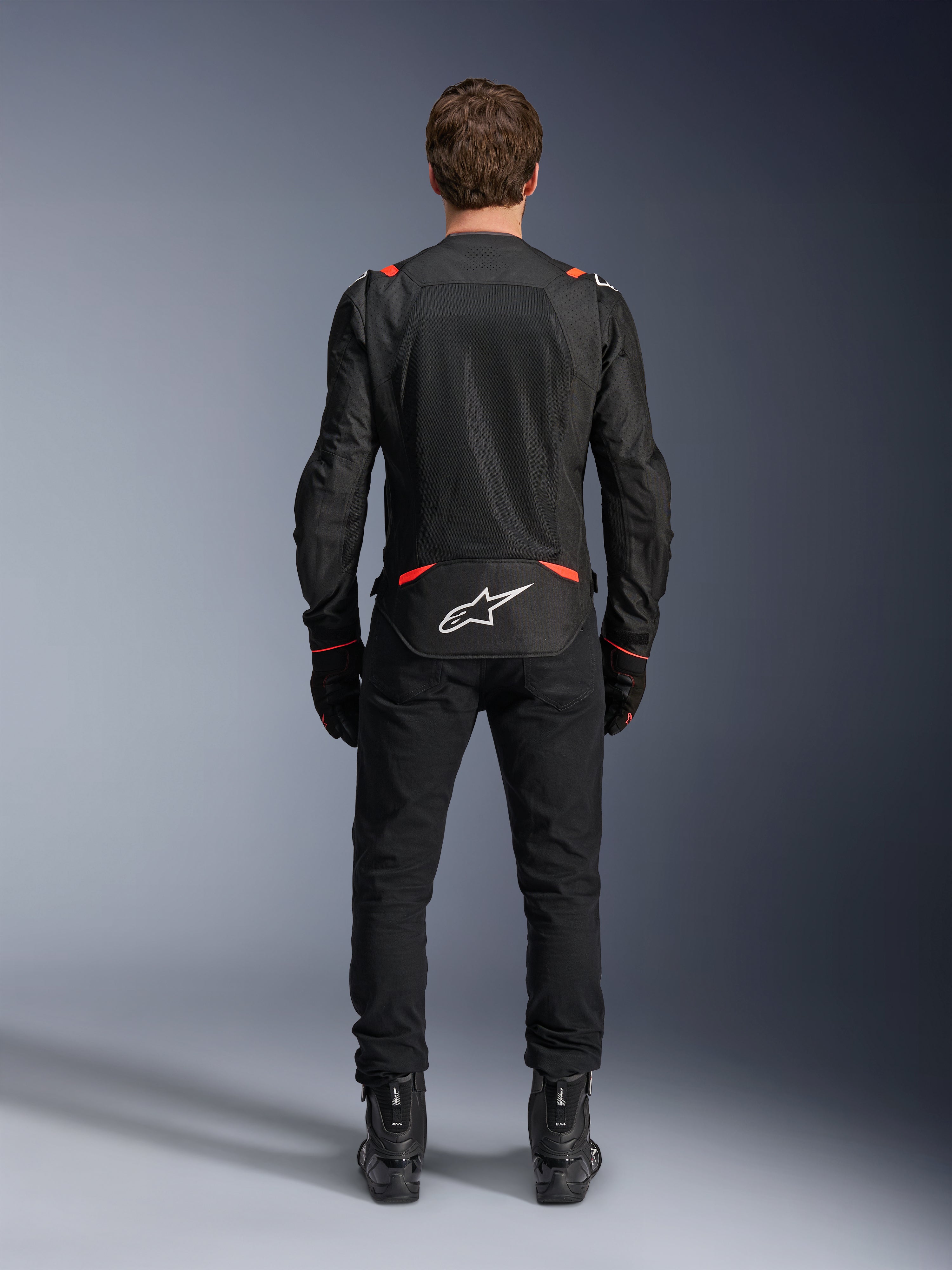 Giacca Alpinestars T-SPS Air v2 in Nero e Rosso Fluo, vista posteriore che mostra ampi pannelli di ventilazione in rete e il logo Alpinestars bianco sulla parte bassa della schiena, indossata con pantaloni da guida e stivali neri.