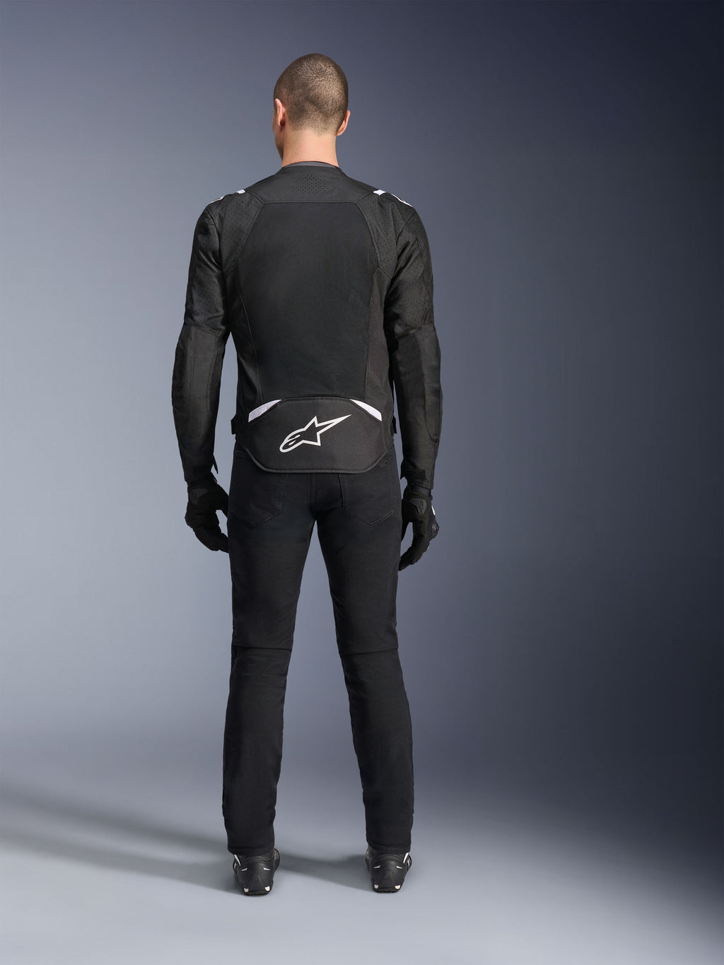 Giacca Alpinestars T-Sps Air V2, vista posteriore, giacca da moto in tessuto nero e bianco, dotata di un ampio pannello posteriore in mesh per il massimo flusso d'aria e logo Astars bianco sulla parte bassa della schiena, indossata con pantaloni e guanti da moto neri