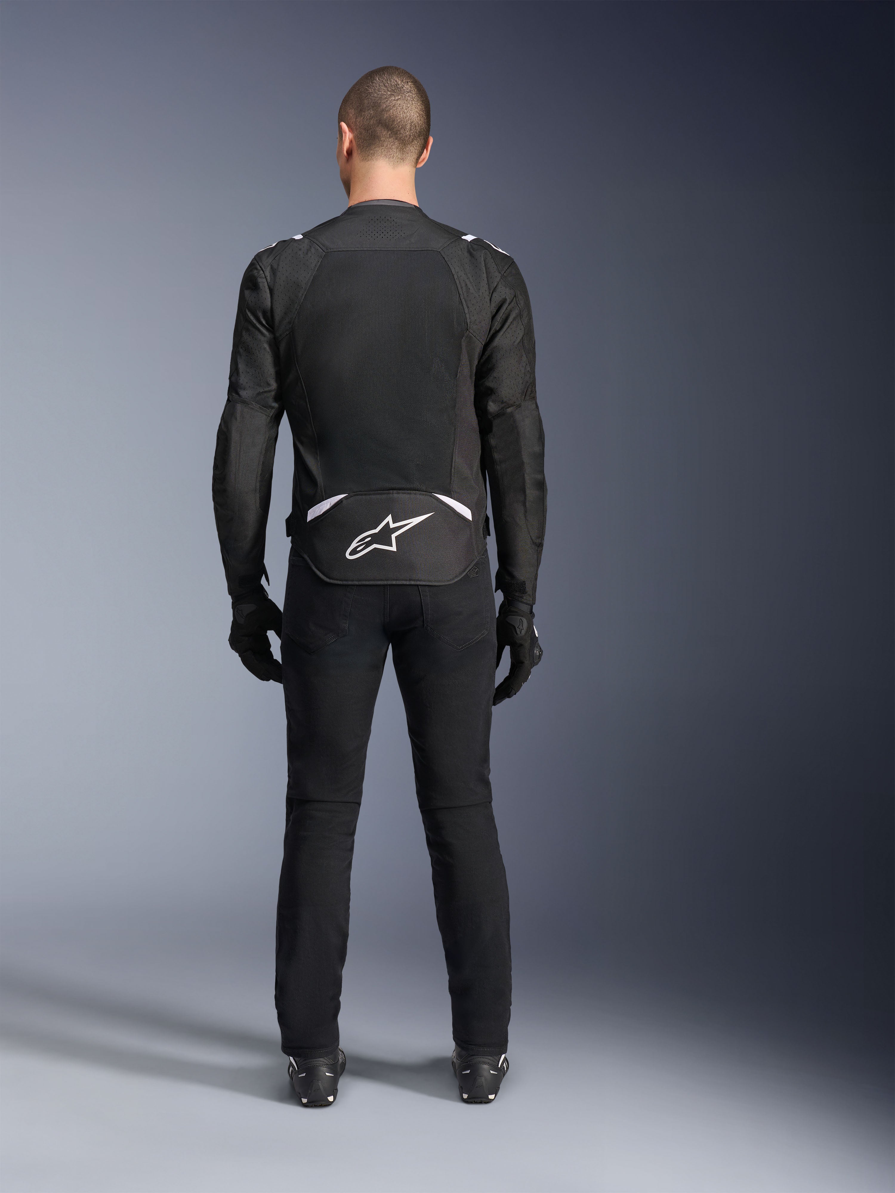 Giacca Alpinestars T-Sps Air V2, vista posteriore, giacca da moto in tessuto nero e bianco, dotata di un ampio pannello posteriore in mesh per il massimo flusso d'aria e logo Astars bianco sulla parte bassa della schiena, indossata con pantaloni e guanti da moto neri