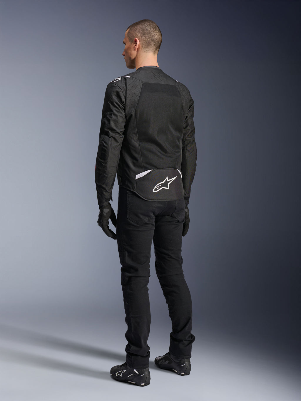 Giacca Alpinestars T-Sps Air V2, giacca da moto in tessuto, nera, vista posteriore con ampio pannello di ventilazione in mesh e logo Alpinestars bianco sulla parte bassa della schiena, abbinata a pantaloni e guanti neri per un look urbano sportivo.
