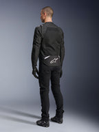 Giacca Alpinestars T-Sps Air V2, giacca da moto in tessuto, nera, vista posteriore con ampio pannello di ventilazione in mesh e logo Alpinestars bianco sulla parte bassa della schiena, abbinata a pantaloni e guanti neri per un look urbano sportivo.
