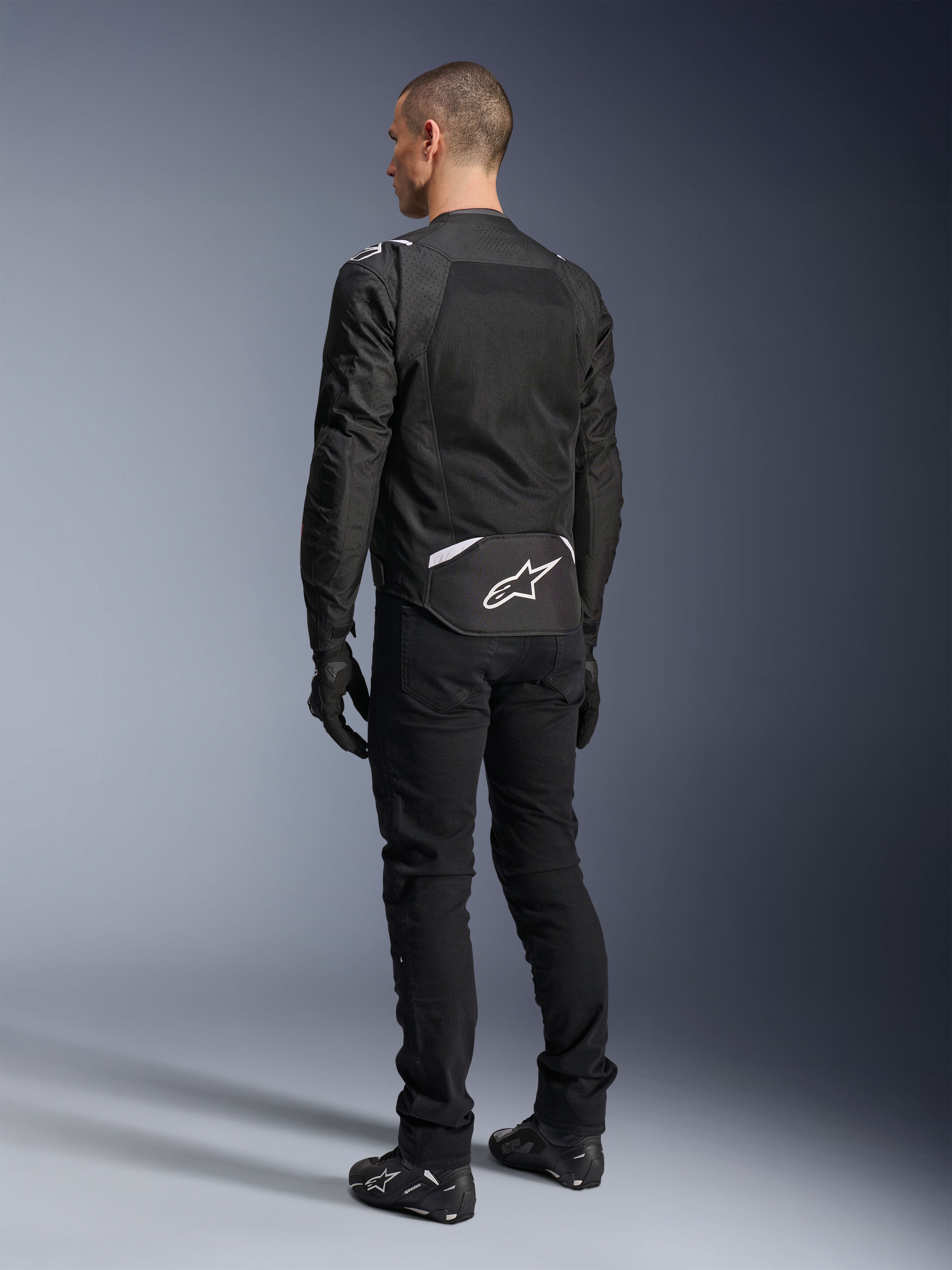 Giacca Alpinestars T-Sps Air V2, giacca da moto in tessuto, nera, vista posteriore con ampio pannello di ventilazione in mesh e logo Alpinestars bianco sulla parte bassa della schiena, abbinata a pantaloni e guanti neri per un look urbano sportivo.